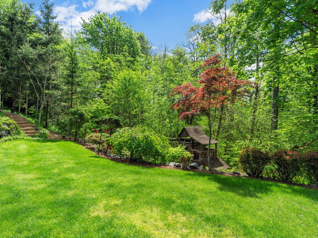 A lush green backyard with a manicured lawn, surrounded by dense trees and bushes. A small wooden gazebo nestles among the plants. A stone pathway leads up the hillside, bordered by vibrant red and green foliage, under a clear blue sky.