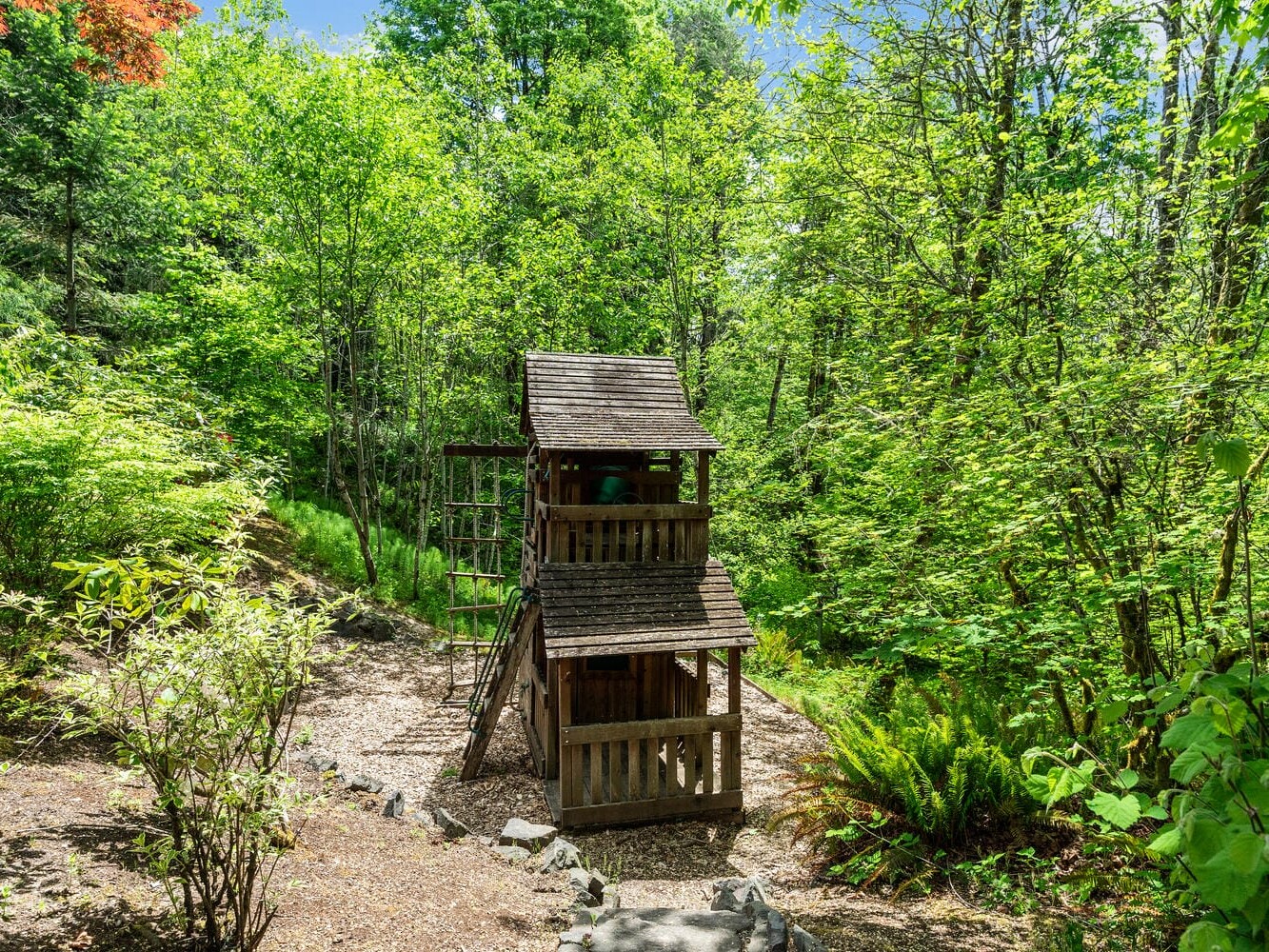 A wooden playhouse with multiple levels is nestled in a lush, green forest. The surrounding area is covered in dense foliage and trees, with a clear path leading to the structure. Sunlight filters through the leaves, creating a serene and inviting atmosphere.