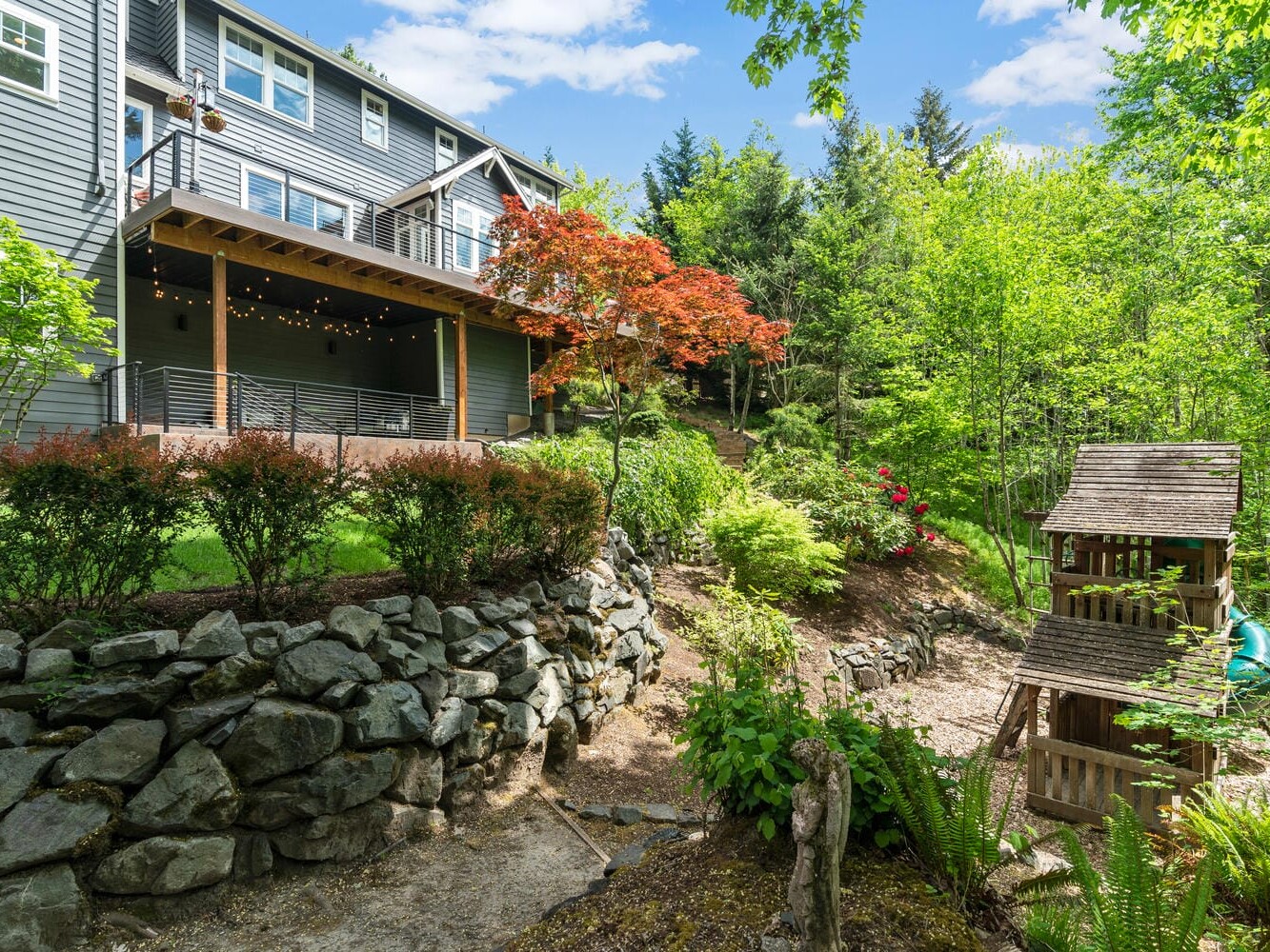A backyard featuring a gray house with a deck adorned with string lights. The garden includes a small wooden playhouse, a stone retaining wall, lush green trees, and a red-leaved tree. A patch of blue sky and clouds is visible above.