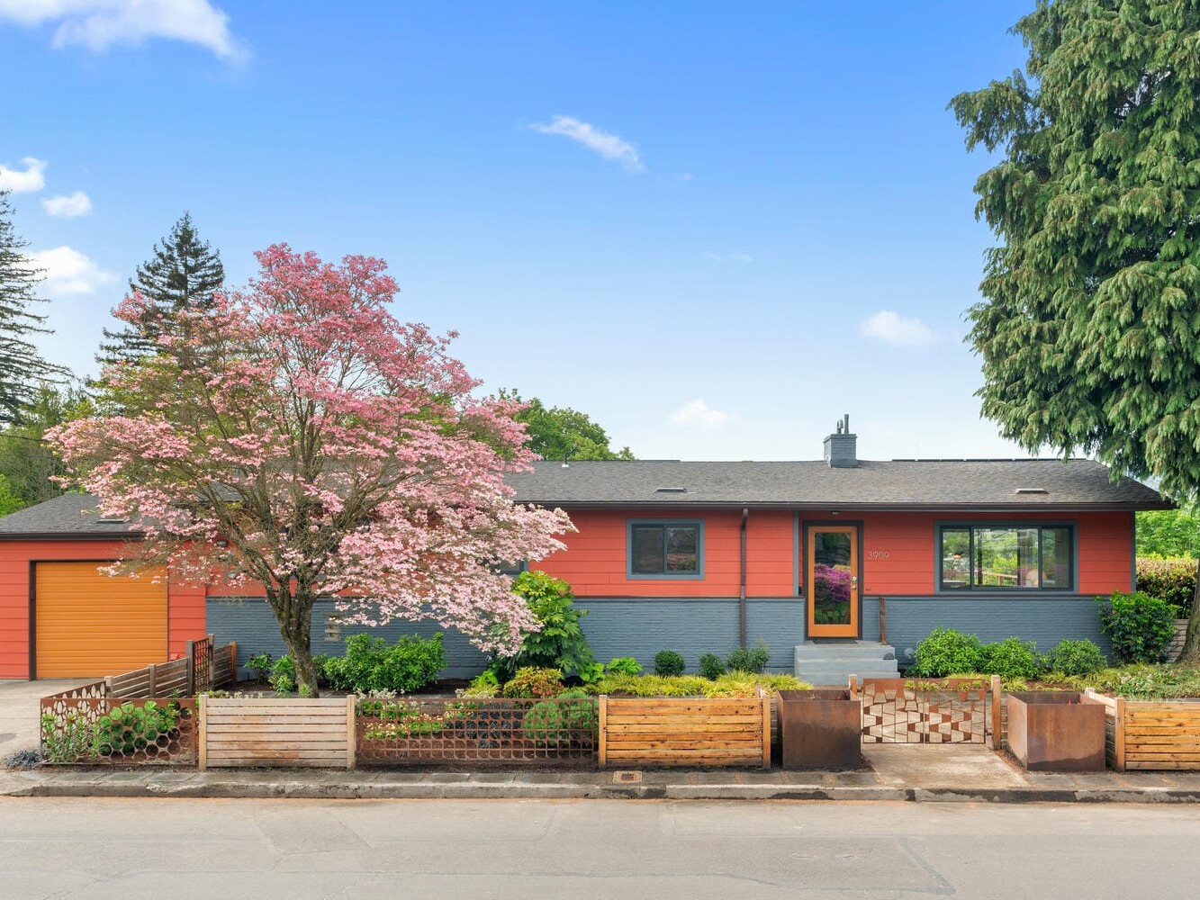 A mid-century modern house with an orange and gray exterior, and a black roof. The front yard features a large pink flowering tree and wooden fencing, with lush greenery and a tall tree on the right. The sky is clear and blue.