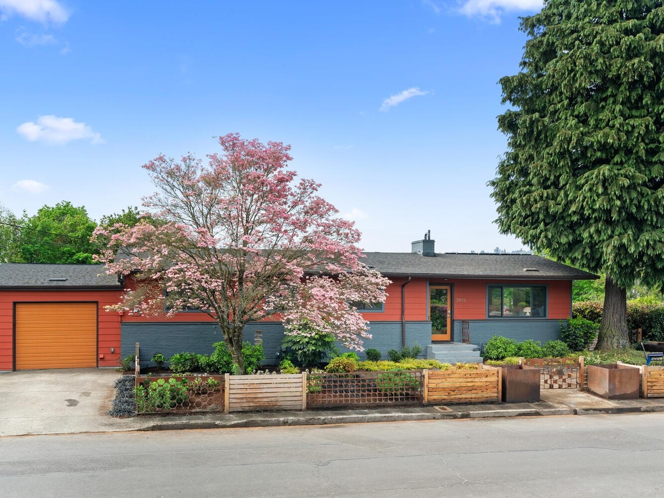 A single-story house with a red and gray exterior, featuring a large pink flowering tree and a wooden fence in the front yard. There are a matching garage door and a lush green tree on the right. The sky is clear and blue.