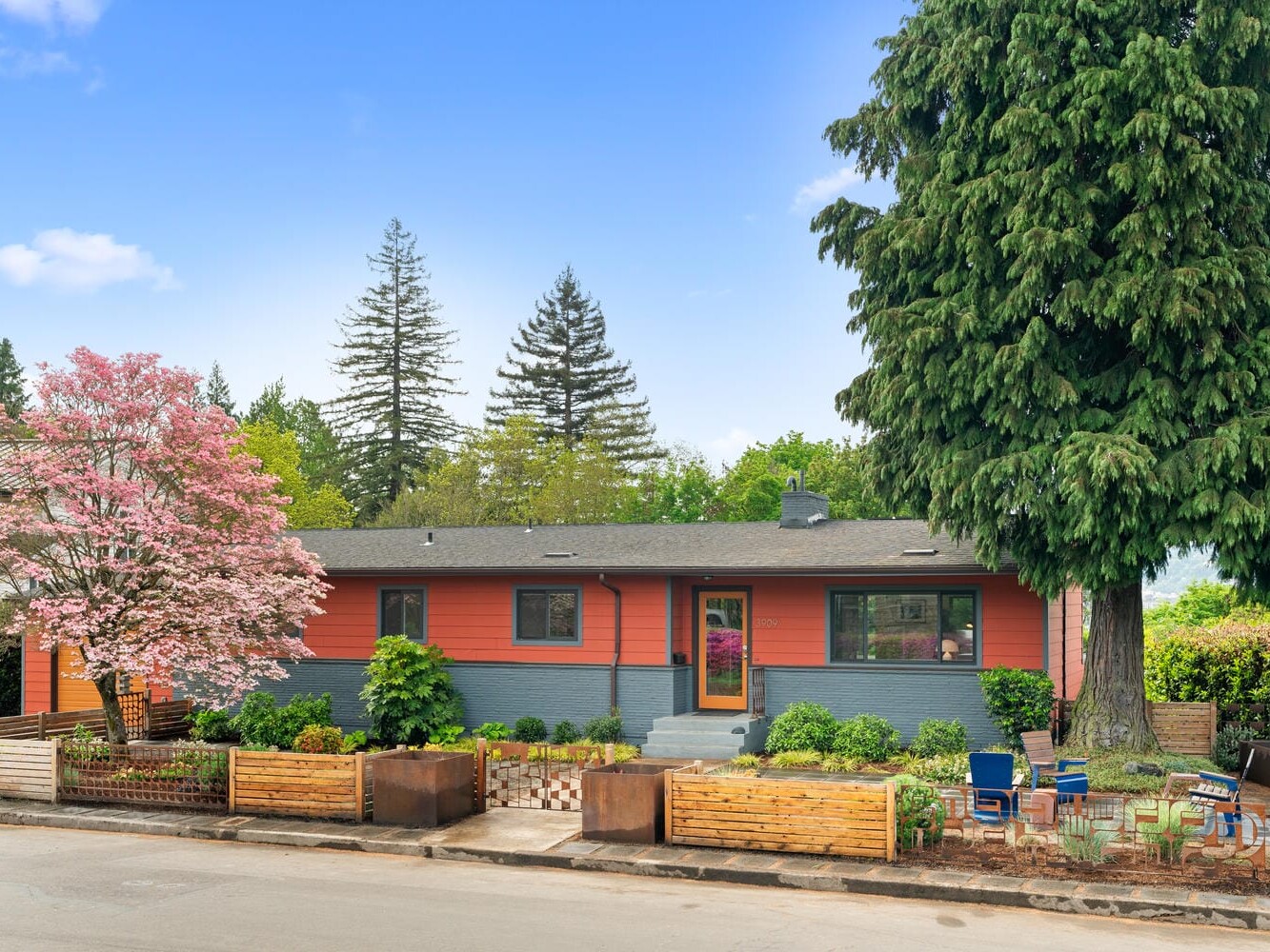 A single-story house with an orange and gray exterior, surrounded by lush trees and a garden. A large pine tree stands on the right, while a pink-blossomed tree is on the left. The front porch has colorful chairs and planters.