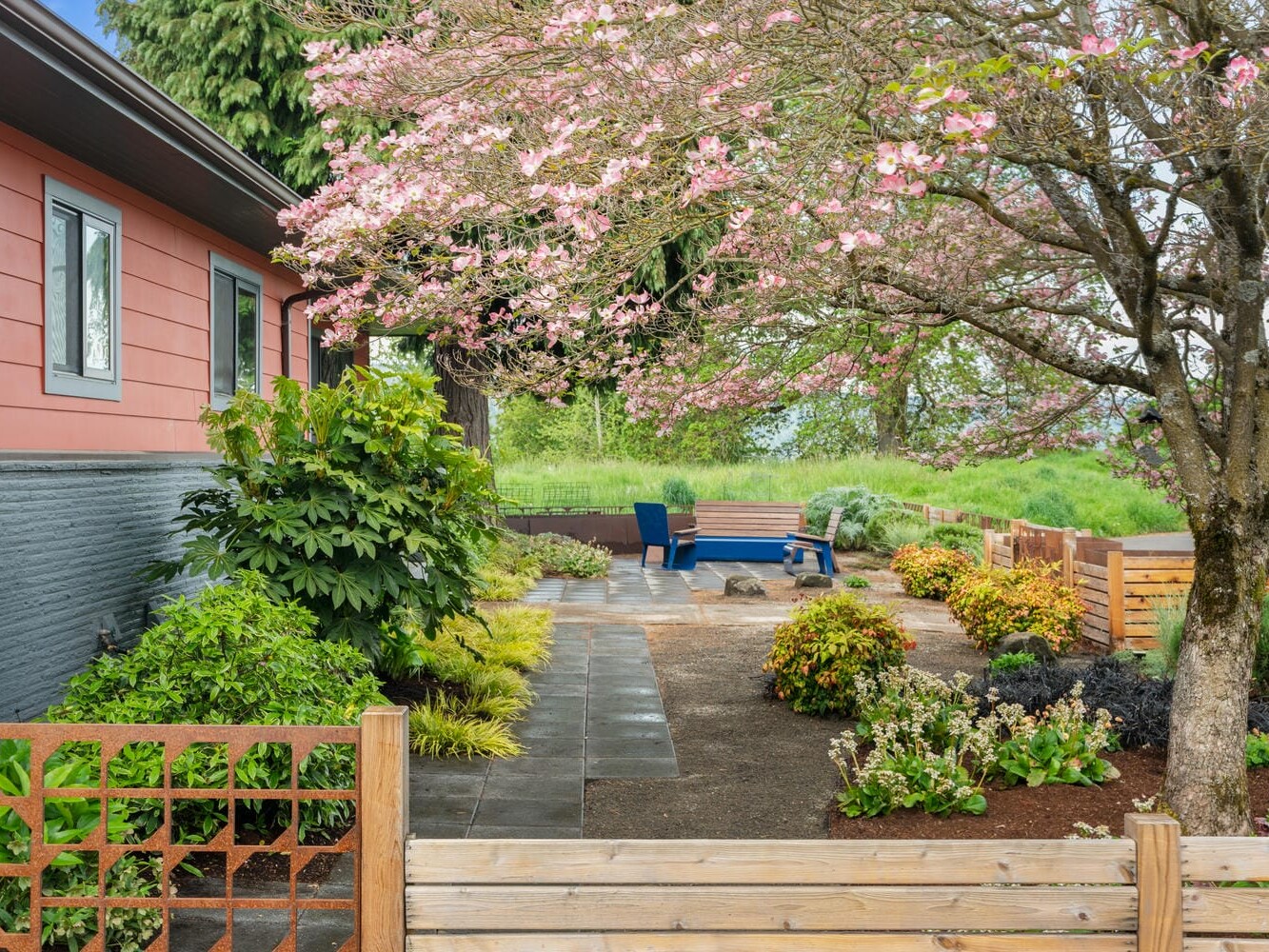 A vibrant garden with a small patio featuring two blue chairs and a table. A cherry blossom tree in full bloom shades the area. The house has red siding and a gray foundation, surrounded by lush greenery and trimmed shrubs. A wooden fence outlines the scene.