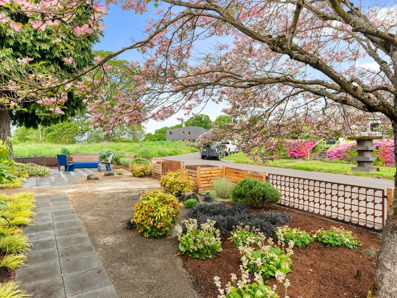 A landscaped garden with a variety of plants and shrubs, bordered by a pathway. A wooden gate opens to a road lined with blooming pink and purple flowers. A tree with pink blossoms provides partial shade on a sunny day.