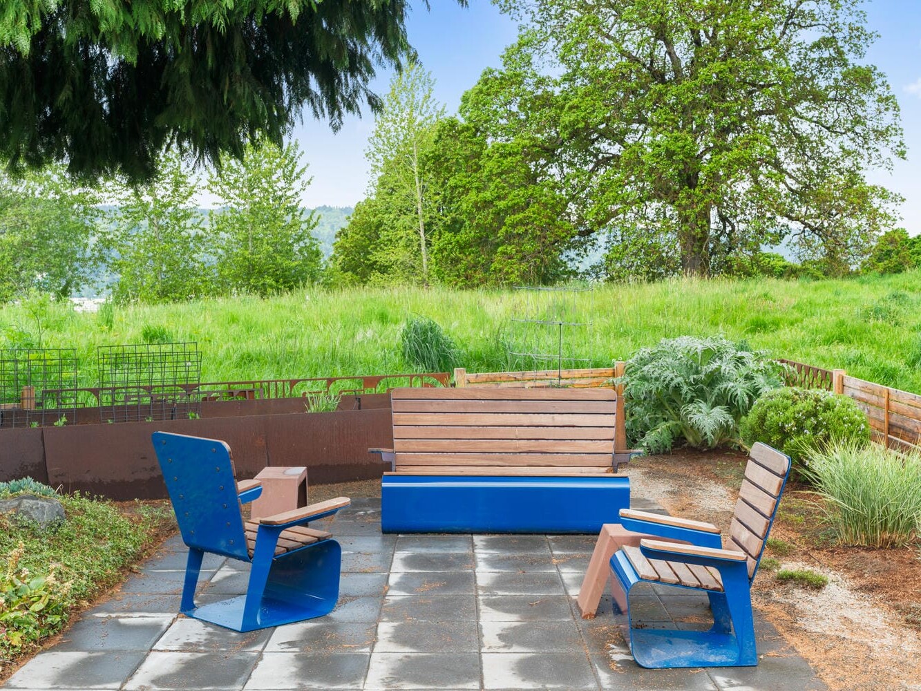 A peaceful garden scene with a central blue-and-wooden bench, two matching blue chairs, and a blue table on a stone patio. The background features lush green grass, trees, and a clear blue sky.