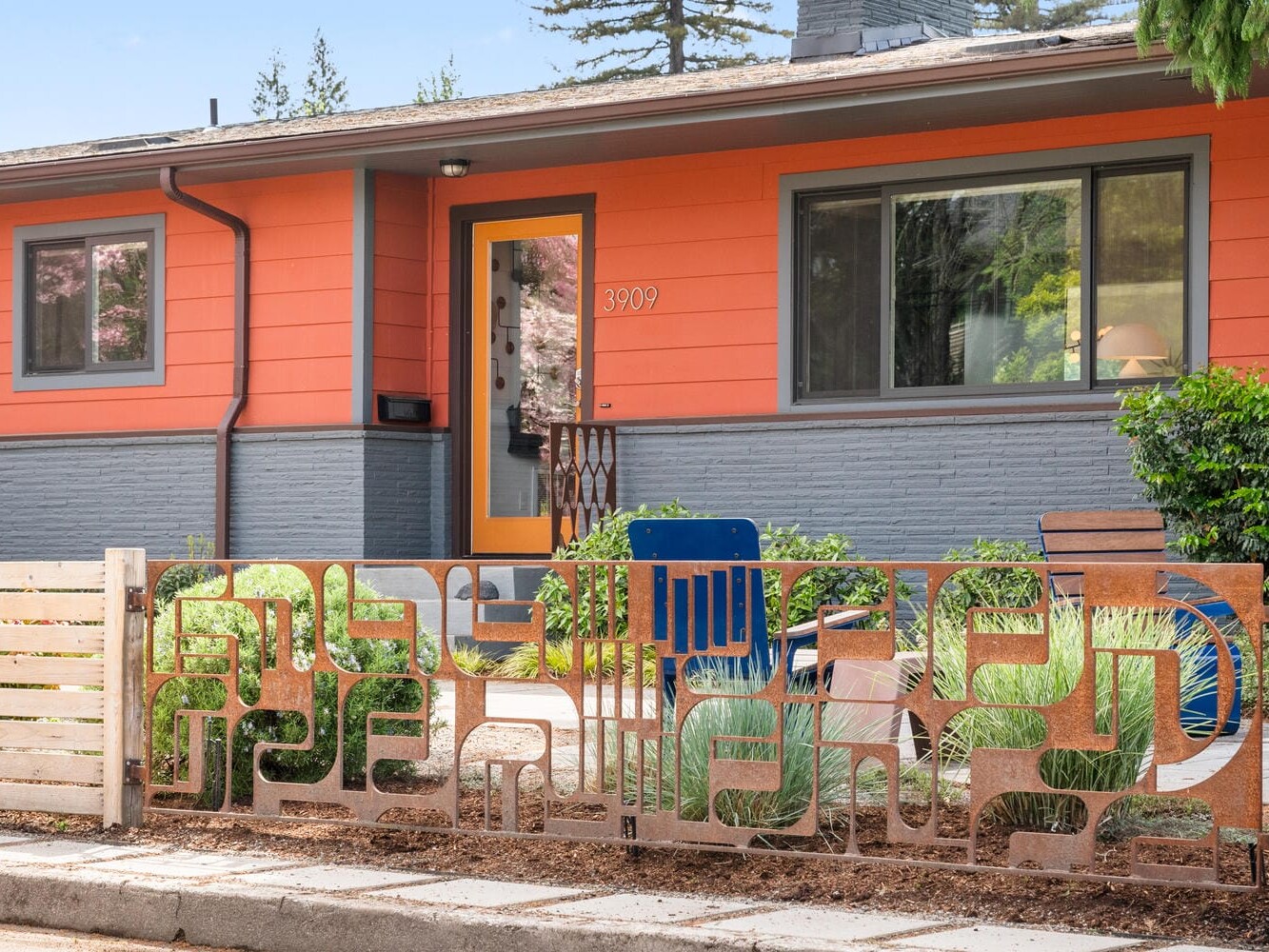 A modern house with an orange and gray exterior features a uniquely designed metal fence with abstract patterns. Theres a glass front door, a blue chair on the porch, and green shrubs in the front yard.