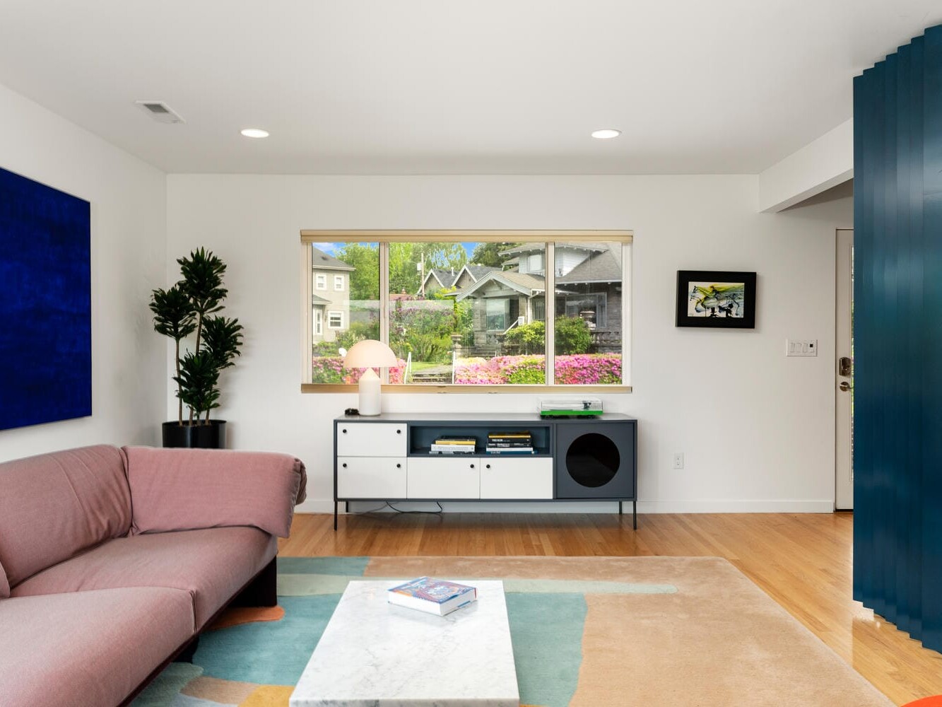 Modern living room with a pink sofa, blue accent wall, and a large blue painting. A window offers a view of a lush garden. The room features a white console table with a lamp and potted plant, wooden flooring, and a marble coffee table.