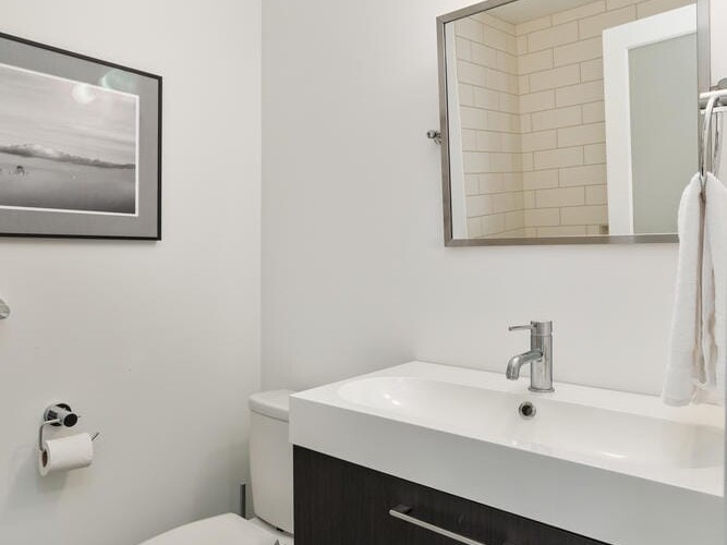 Modern bathroom featuring a white toilet, dark wood vanity with a white sink, and a mirror above. A towel hangs on the right, and a framed photograph is on the light-colored wall. A wall-mounted light fixture illuminates the space.