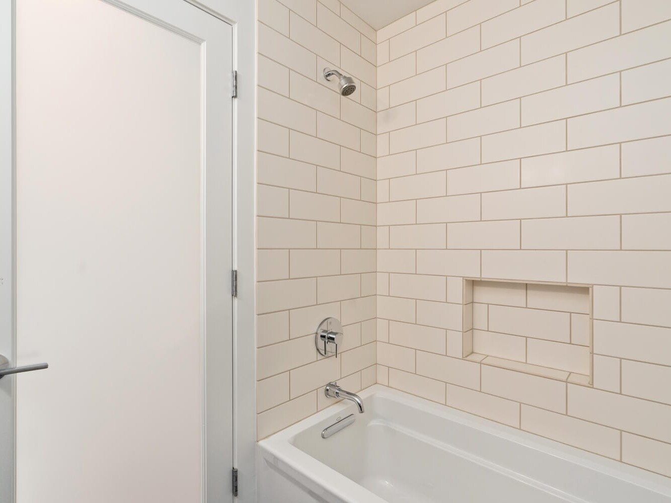 A bathroom showing a white tiled wall with a built-in shelf, a bathtub, and a showerhead. The door to the left has a silver handle. The tiles have a light grout, giving a clean and minimalist appearance.