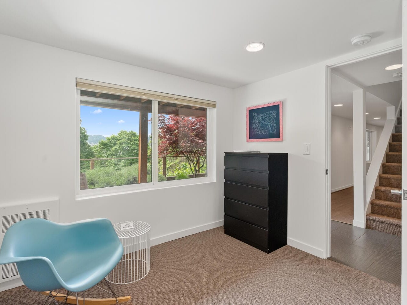 A small, bright room features a large window with scenic outdoor views, a modern blue chair, and a black chest of drawers. A white side table sits beside the chair, and a framed picture is on the wall. A staircase is visible in the background.