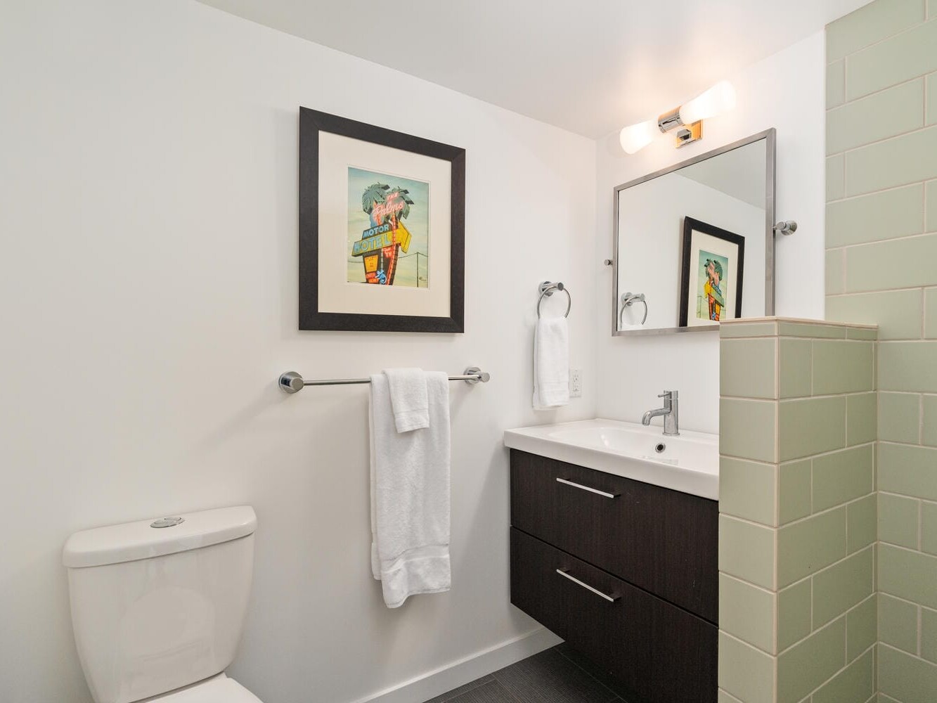 A modern bathroom with white walls features a toilet, a wooden vanity with a mirror and light fixture, and green tile accents. A towel hangs on a rail, and a colorful framed artwork is displayed above.