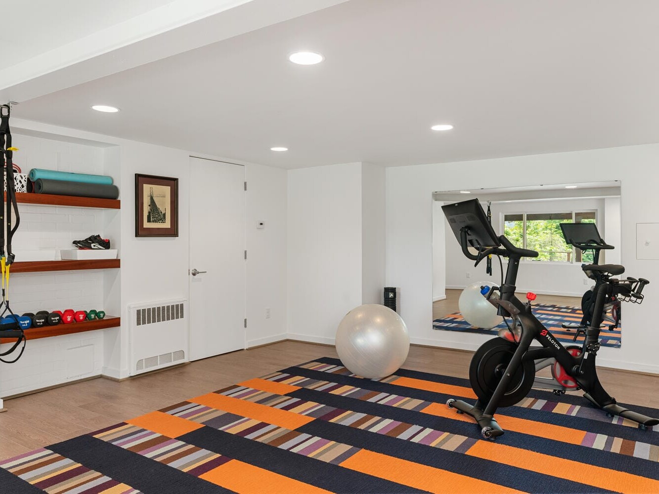 A modern home gym with a stationary bike, exercise ball, and striped orange and purple rug. Shelves hold yoga mats, small dumbbells, and sneakers. A large mirror reflects a window showing greenery. White walls and ceiling lights complete the space.