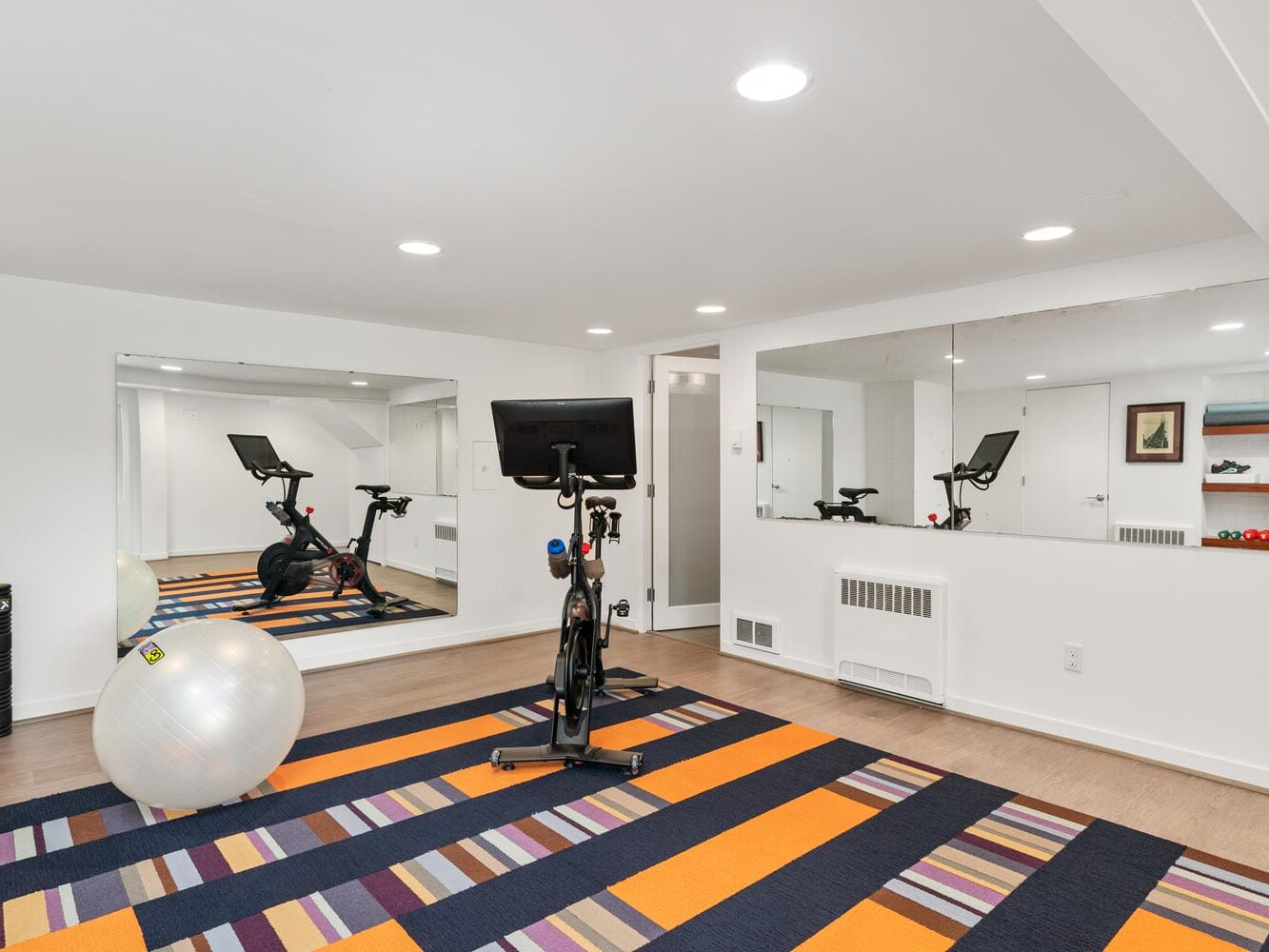 A home gym with a stationary bike, a large exercise ball, and striped orange and purple carpet. Mirrors line the walls, reflecting the equipment. The space is well-lit with recessed lighting.