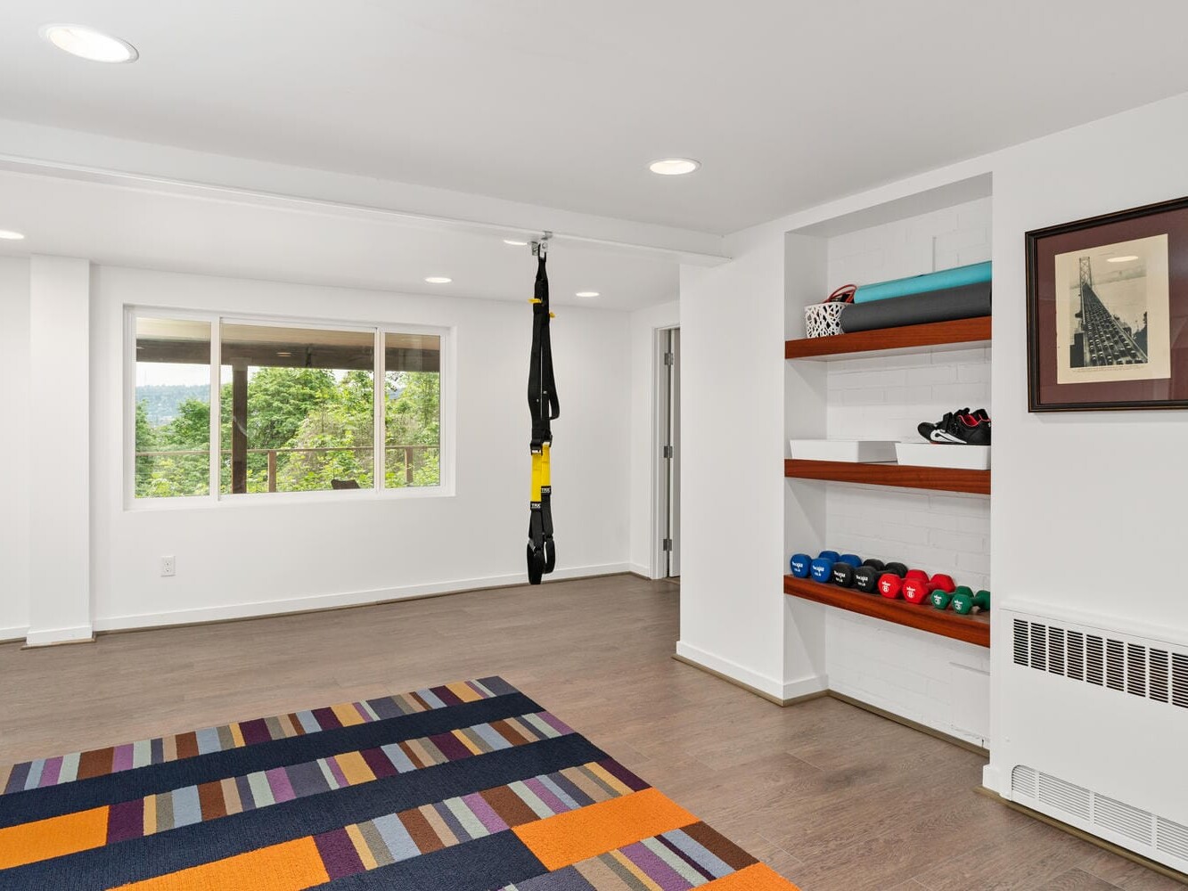 A home gym with a brightly colored striped rug, wall-mounted shelves holding dumbbells, and a suspension trainer hanging from the ceiling. A large window offers a view of greenery outside, and the white walls are adorned with a framed picture.