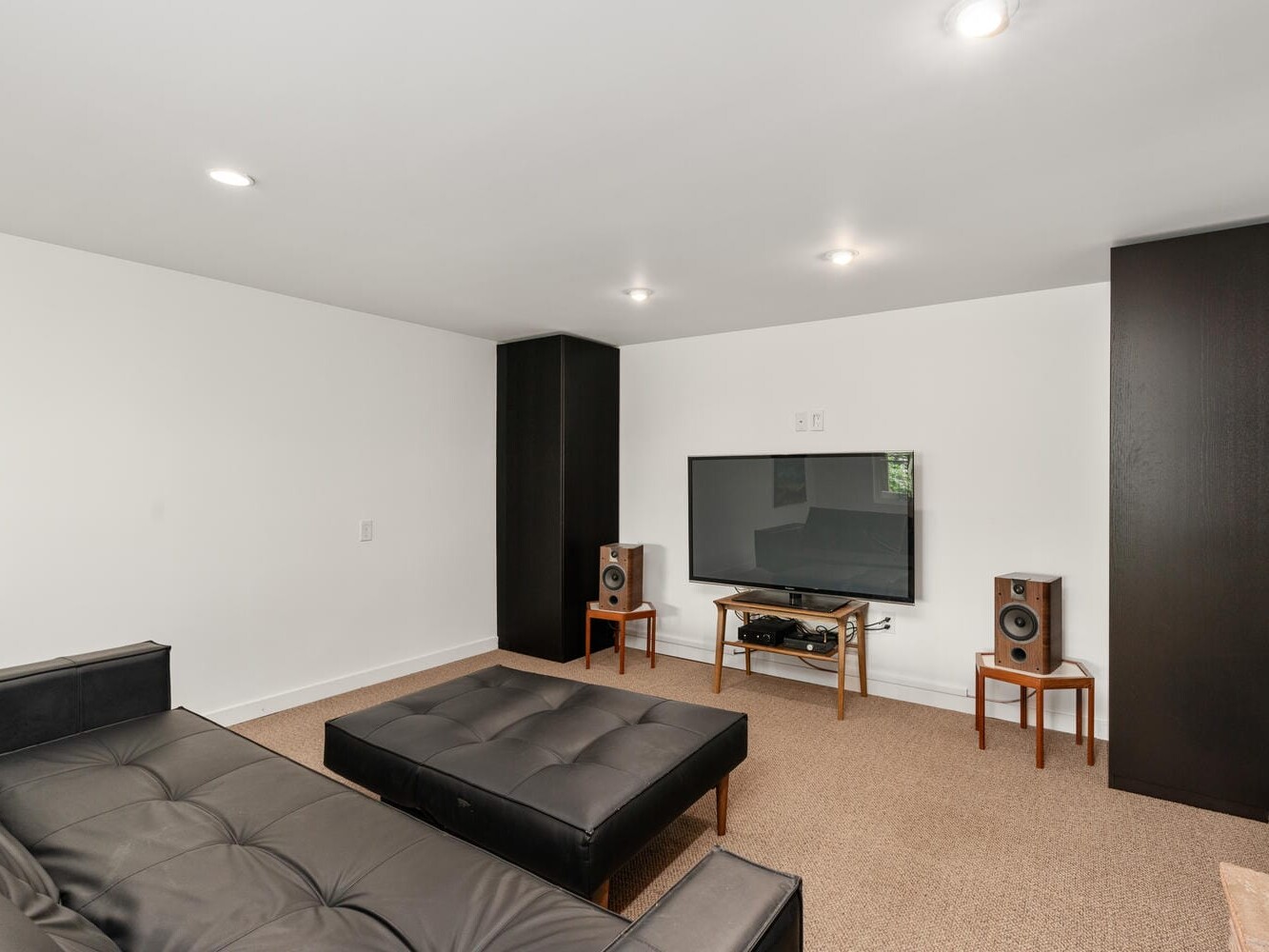 A modern living room with a large flat-screen TV on a wooden stand, flanked by two tall black speakers on chairs. A black leather sectional sofa and ottoman are placed on a carpeted floor. The space is illuminated by recessed lighting.