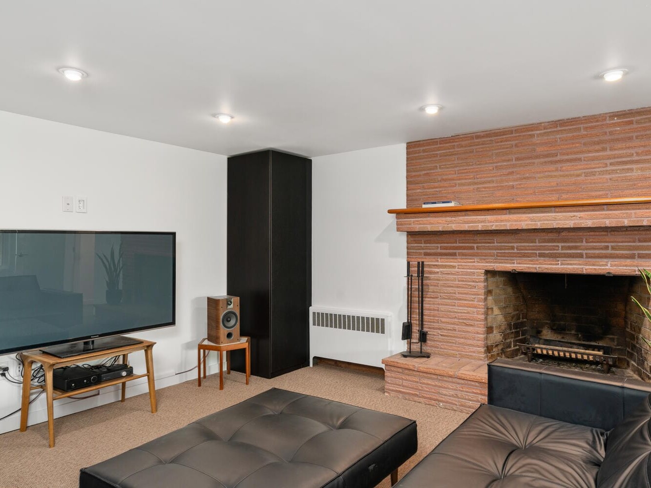 A cozy living room features a mounted flat-screen TV, a black cushioned ottoman, a brick fireplace with a wooden mantle, and a speaker on a wooden stand. The room has white walls, a carpeted floor, and recessed lighting.