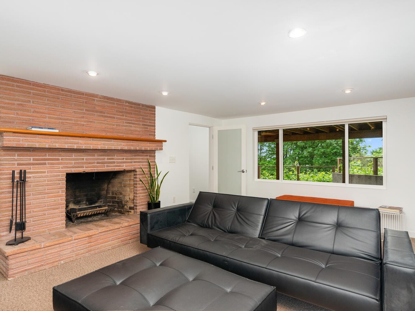 A modern living room with a black leather sofa, ottoman, and a brick fireplace. Theres a large window offering a view of greenery, and a potted plant beside the fireplace. The space is lit by recessed ceiling lights.