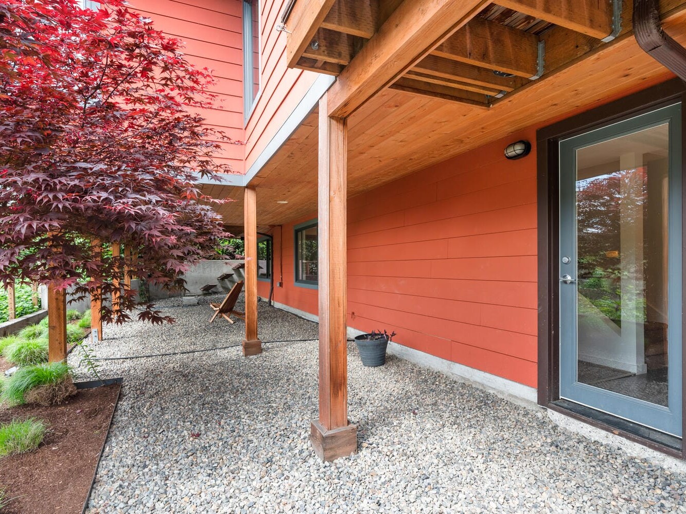 A modern house with red siding features a gravel pathway, wooden support beams, and a glass door. A vibrant red maple tree and greenery are visible beside the walkway. The low overhead structure provides a sheltered outdoor area.