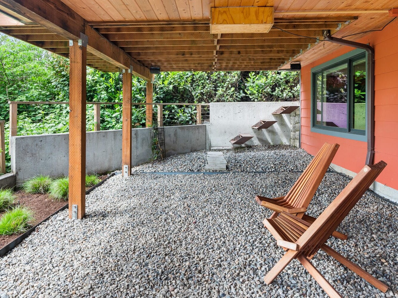 A covered outdoor patio with a gravel floor and two wooden chairs. Theres a red exterior wall with a window on the right, and a small wooden staircase leading to a garden in the background. Flooring includes patches of greenery.