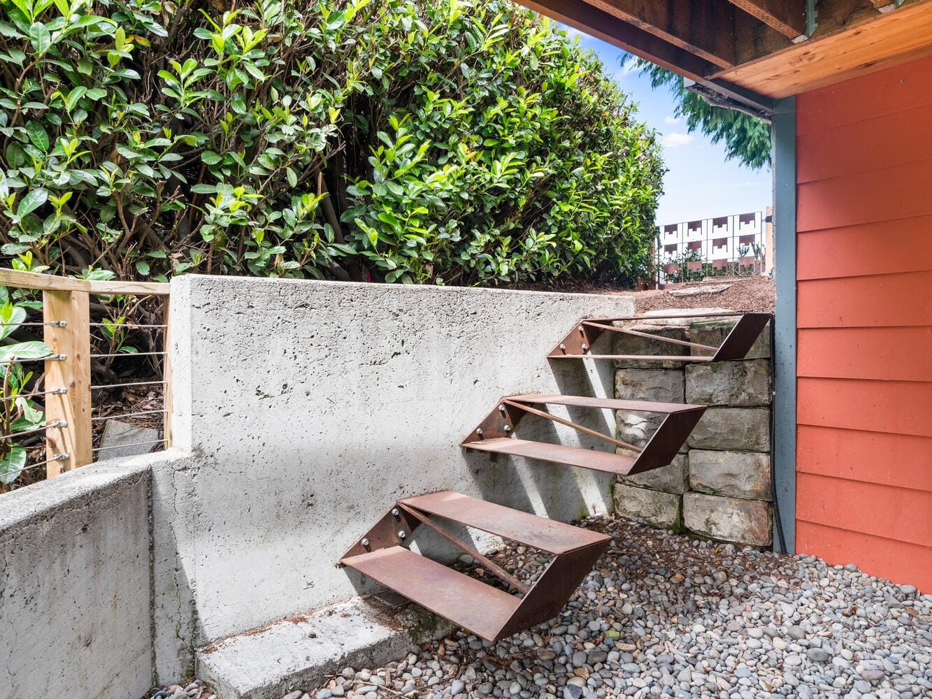 A modern staircase with metal steps leads up to a garden, bordered by lush green hedges. The base is gravel, and a red building with horizontal siding is partially visible. The sky is clear, and apartment buildings can be seen in the background.