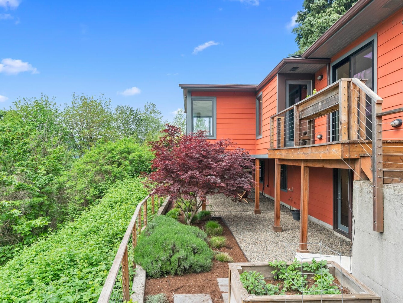 Modern hillside home with vibrant orange exterior. Features wooden railings and balcony overlooking lush greenery. A small garden with shrubs and a Japanese maple add charm. The sky is clear and blue.