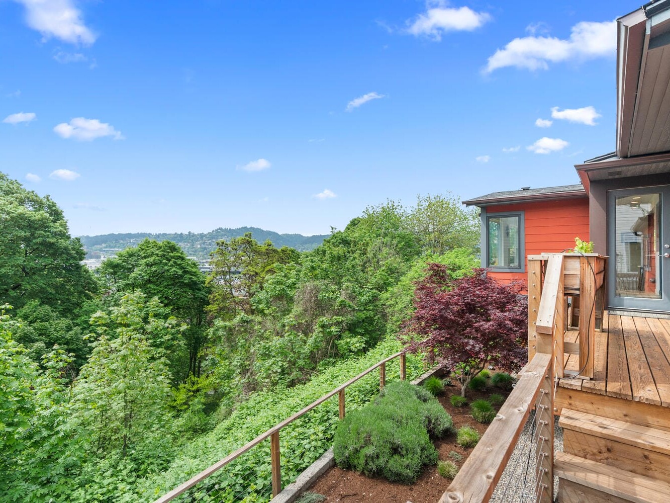 A house with a wooden deck overlooks a lush green landscape, with a forested area extending to distant hills. The sky is clear and blue, creating a serene and picturesque view.