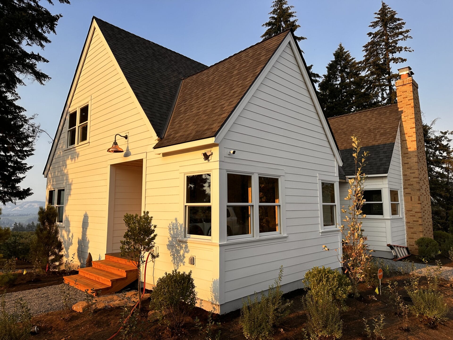 A charming white house with a steep gable roof and chimney is surrounded by trees and shrubs. The sun casts a warm glow on the exterior and the steps leading to the entrance.