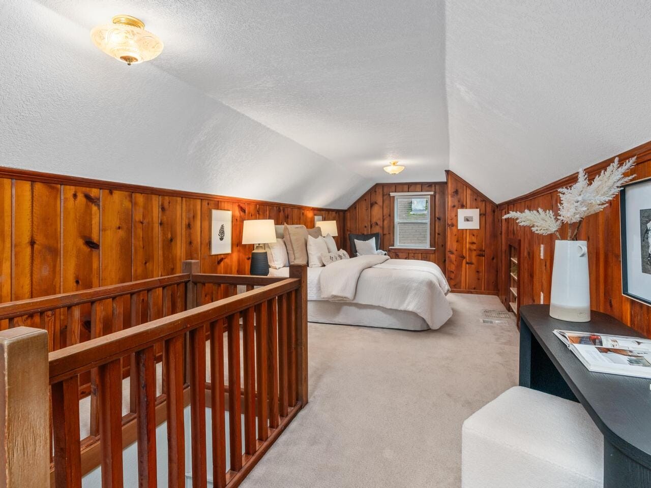 A cozy attic bedroom with wood-paneled walls and sloped ceiling. It features a bed with white linens, a nightstand with a lamp, and a small desk with a vase and decorations. The space is warmly lit and carpeted.