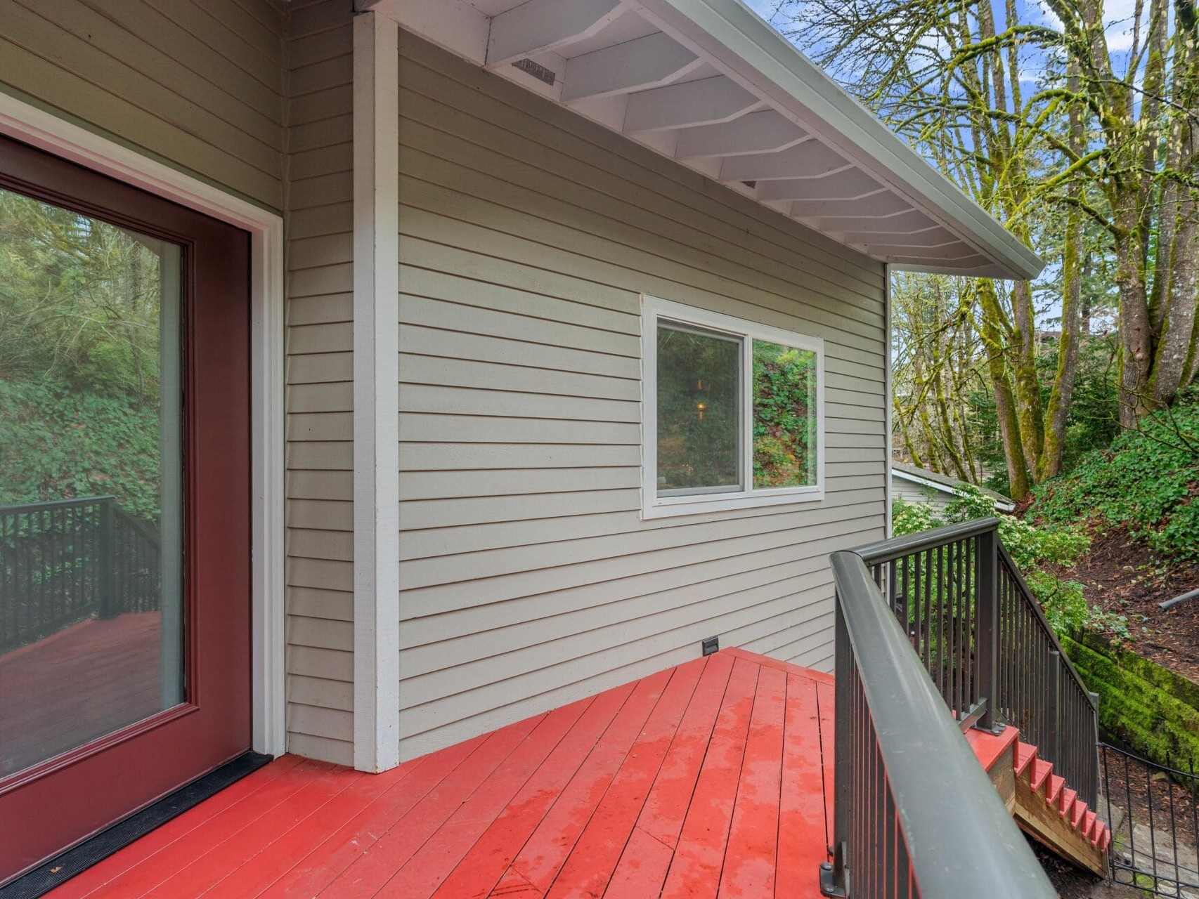 A small deck with red wooden flooring and gray siding on a house. It leads to a glass door on the left and a window in the center. The deck is bordered by a black railing and nearby trees provide a lush, green backdrop.