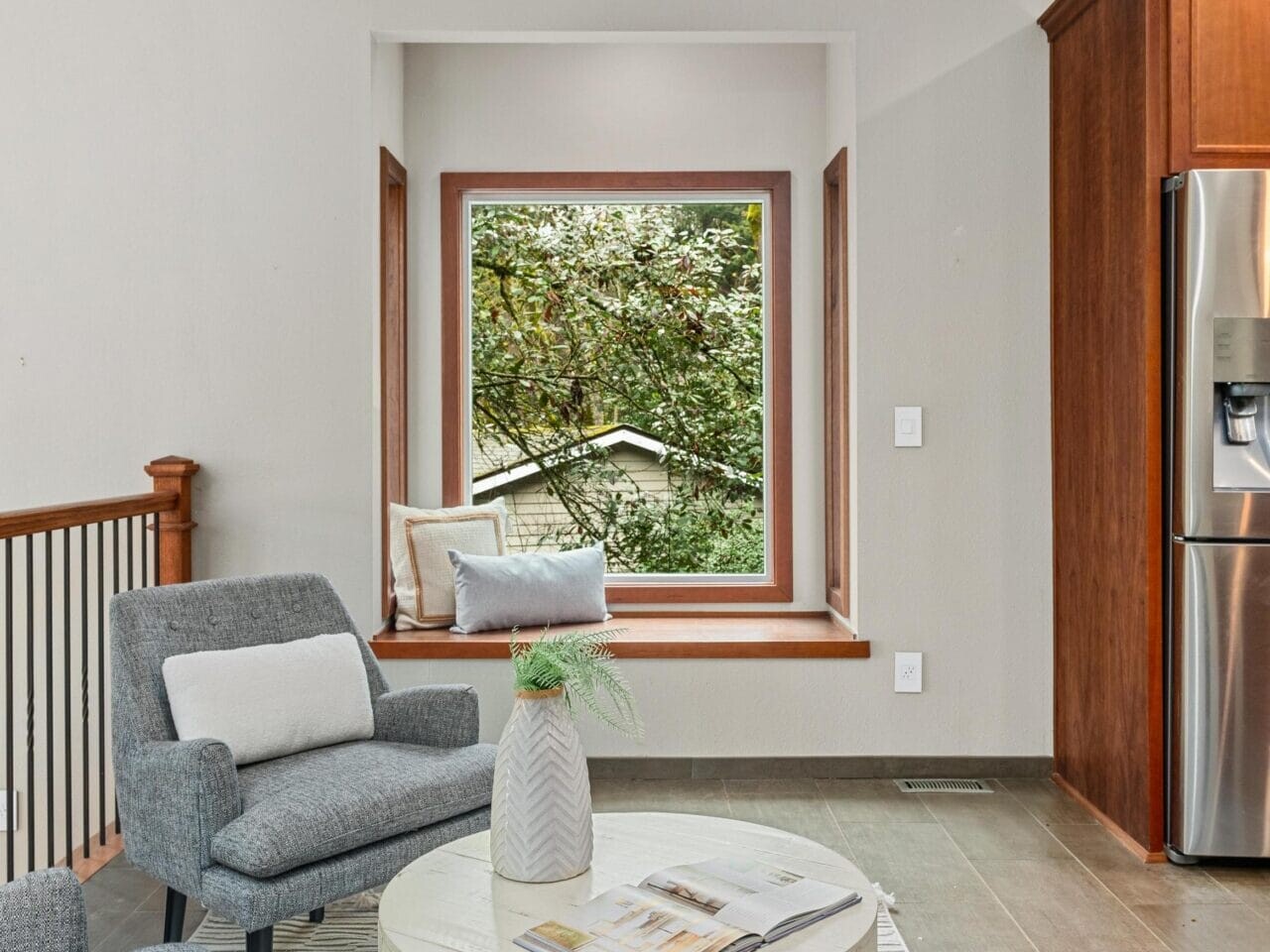 Cozy room with a gray armchair and plaid pillows next to a round white table. A bay window with a view of greenery adds natural light. A beige pendant lamp hangs overhead. Adjacent is a stainless steel refrigerator and dark wooden cabinetry.