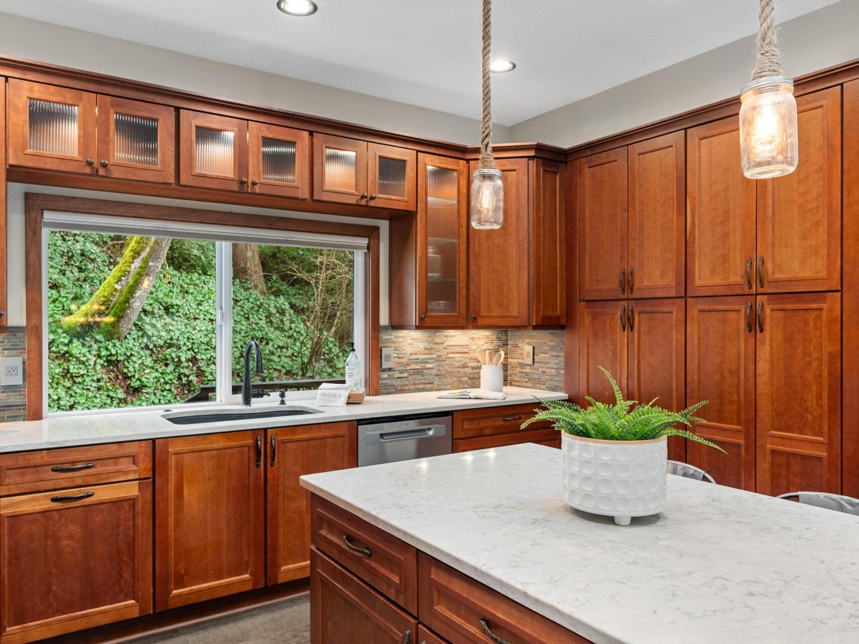 A modern kitchen featuring wooden cabinets with glass accents, a large window overlooking greenery, and a marble island with a potted plant. Pendant lights hang above the island, and there are stainless steel appliances and a black sink.