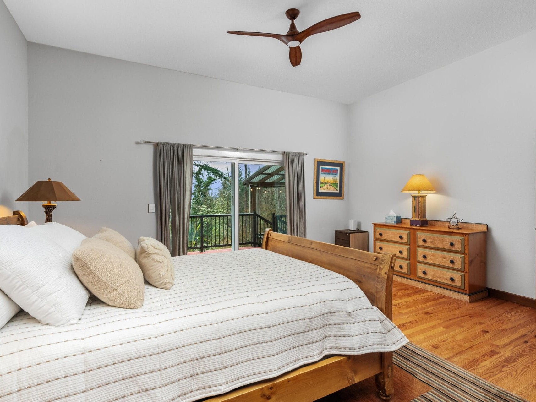 A cozy bedroom with a large wooden bed, white bedding, and beige pillows. Theres a wooden dresser with a lamp and a painting on the wall. A ceiling fan is above, and sliding doors open to a scenic balcony with greenery visible outside.