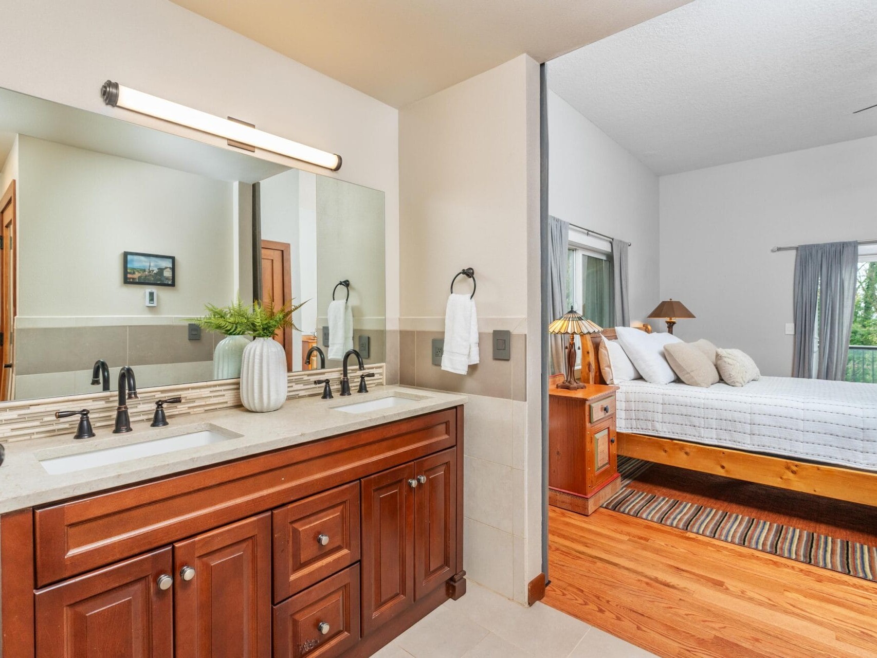 A bathroom with a wooden double vanity, two mirrors, and a modern light fixture. To the side, an open doorway leads to a bedroom with a neatly made bed, wooden floors, and a rug. A vase with greenery decorates the vanity.