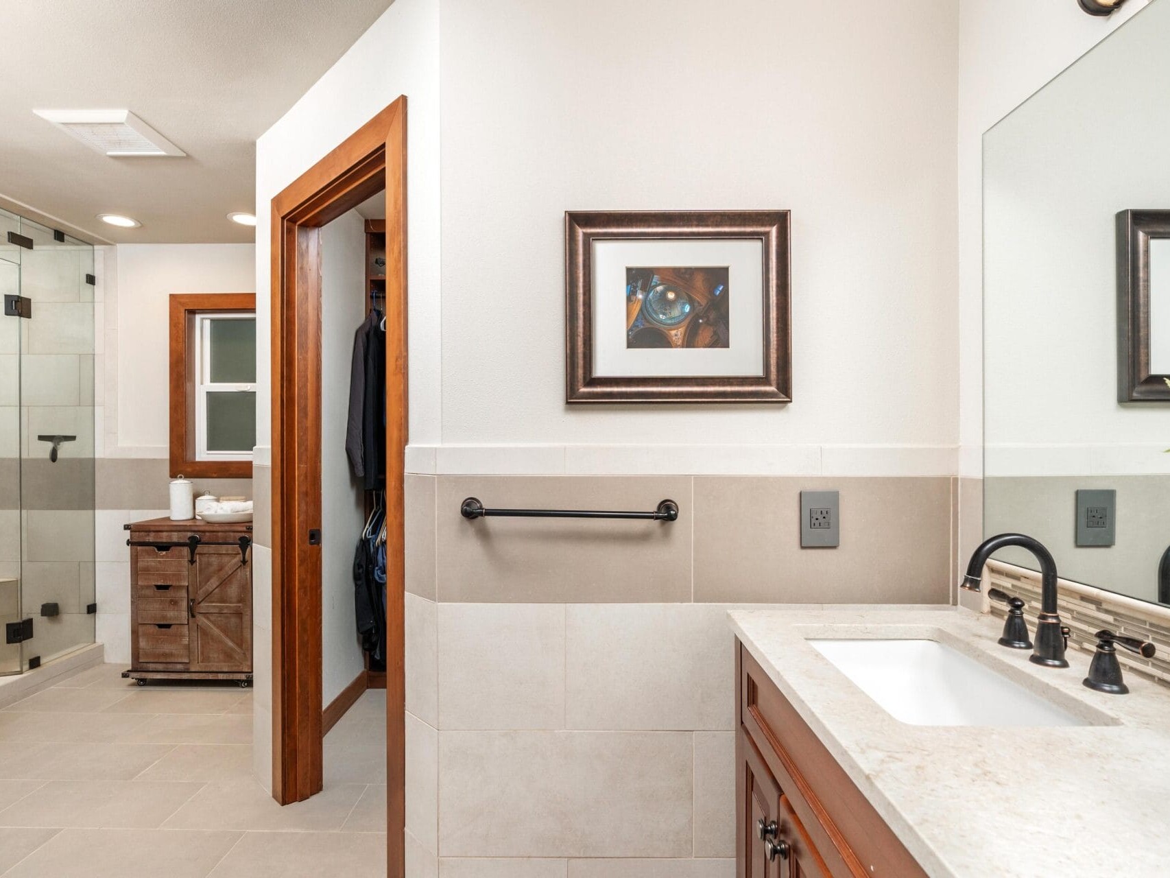 A bathroom with a large mirror, sink, and dark faucet. Theres a towel rack beneath a framed picture. In the background, a walk-in shower with a glass enclosure is visible, along with a wooden cabinet holding towels.