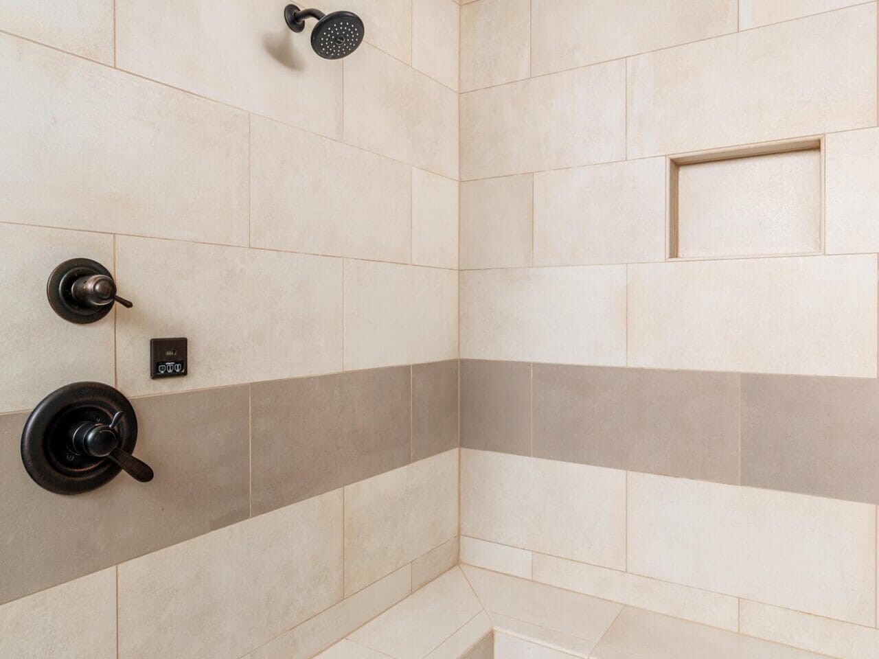 A modern shower with beige tiles, a built-in bench, and a small niche for toiletries. The shower features a black showerhead and controls, with a pebble stone floor adding texture to the design.