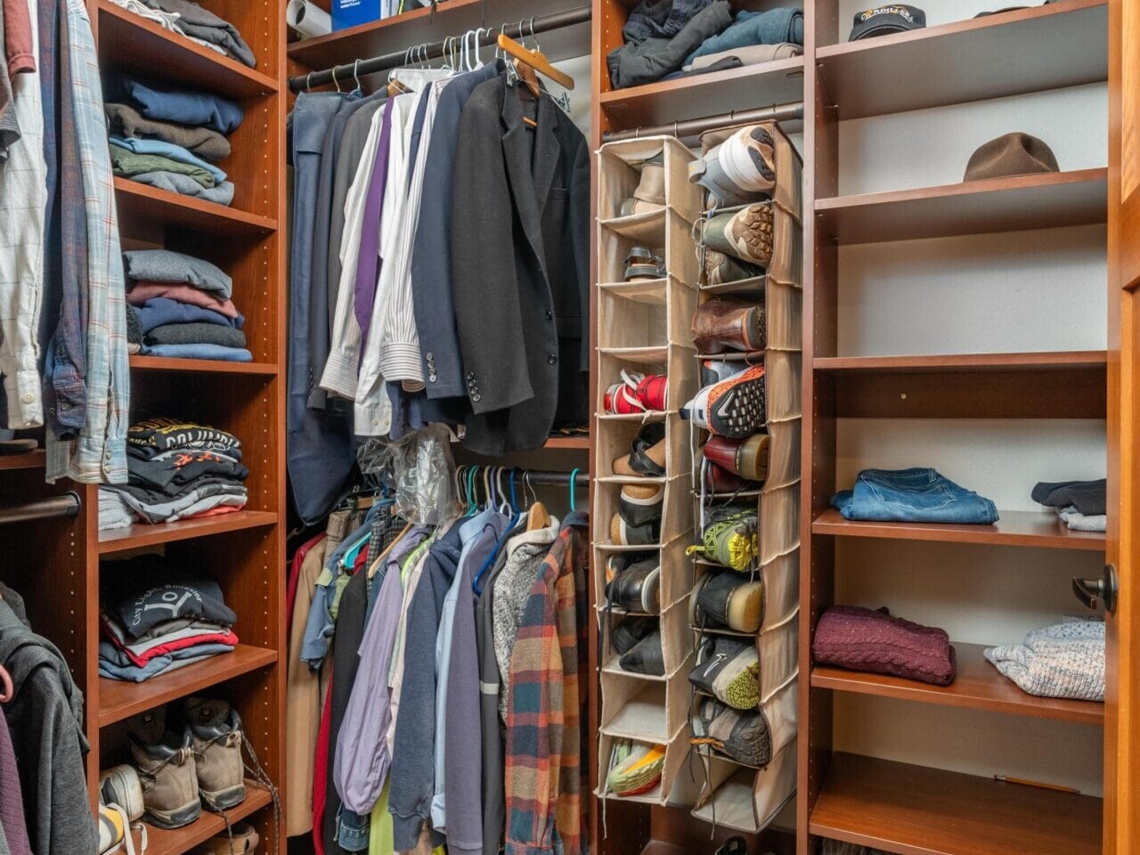 A walk-in closet with wooden shelves filled with neatly arranged clothes, jackets, and shoes. Several shirts and jackets hang on a rod, while shoes and folded clothes are organized on the shelves.