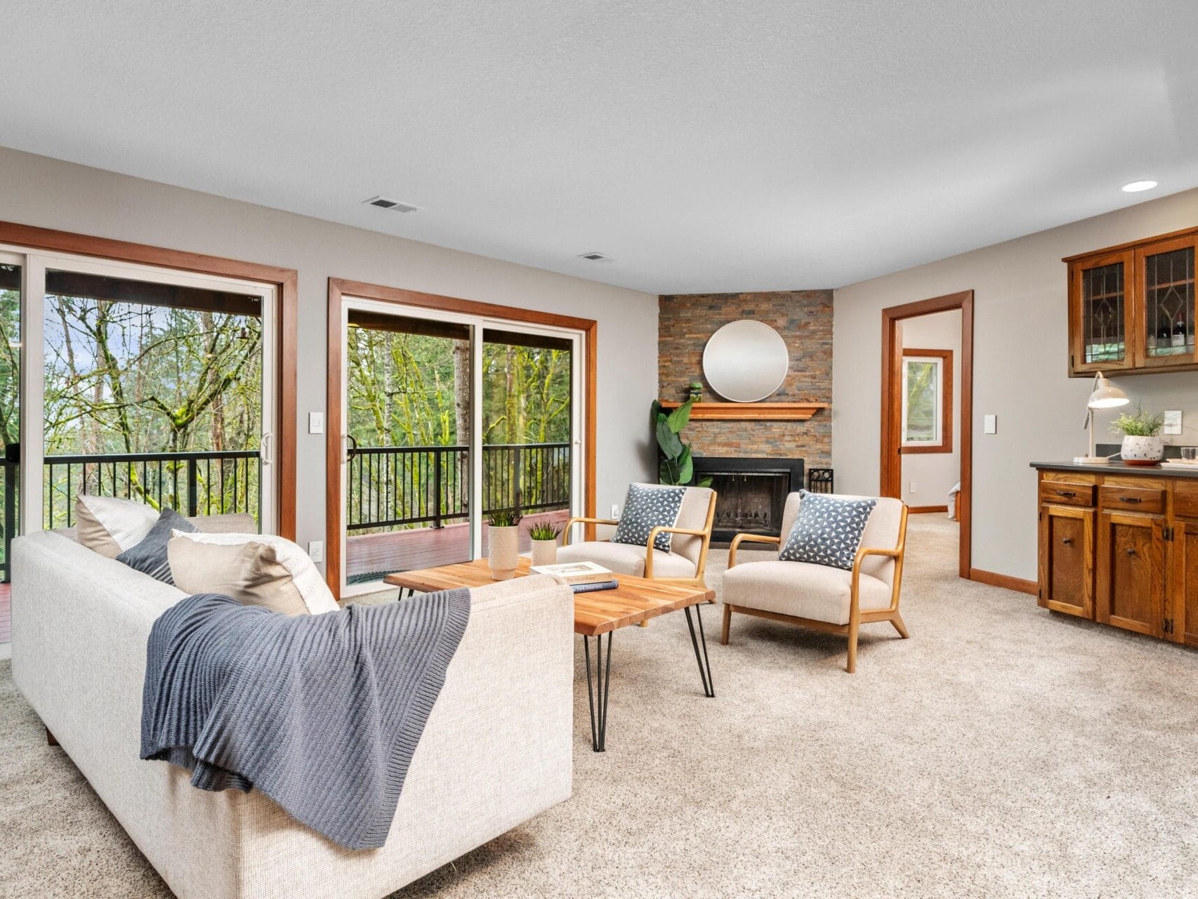 A cozy living room with large windows showing a forest view. Theres a beige sofa with a gray throw, two wooden chairs, a wooden coffee table, a fireplace, and a round mirror above. A wooden cabinet and a plant are in the background.