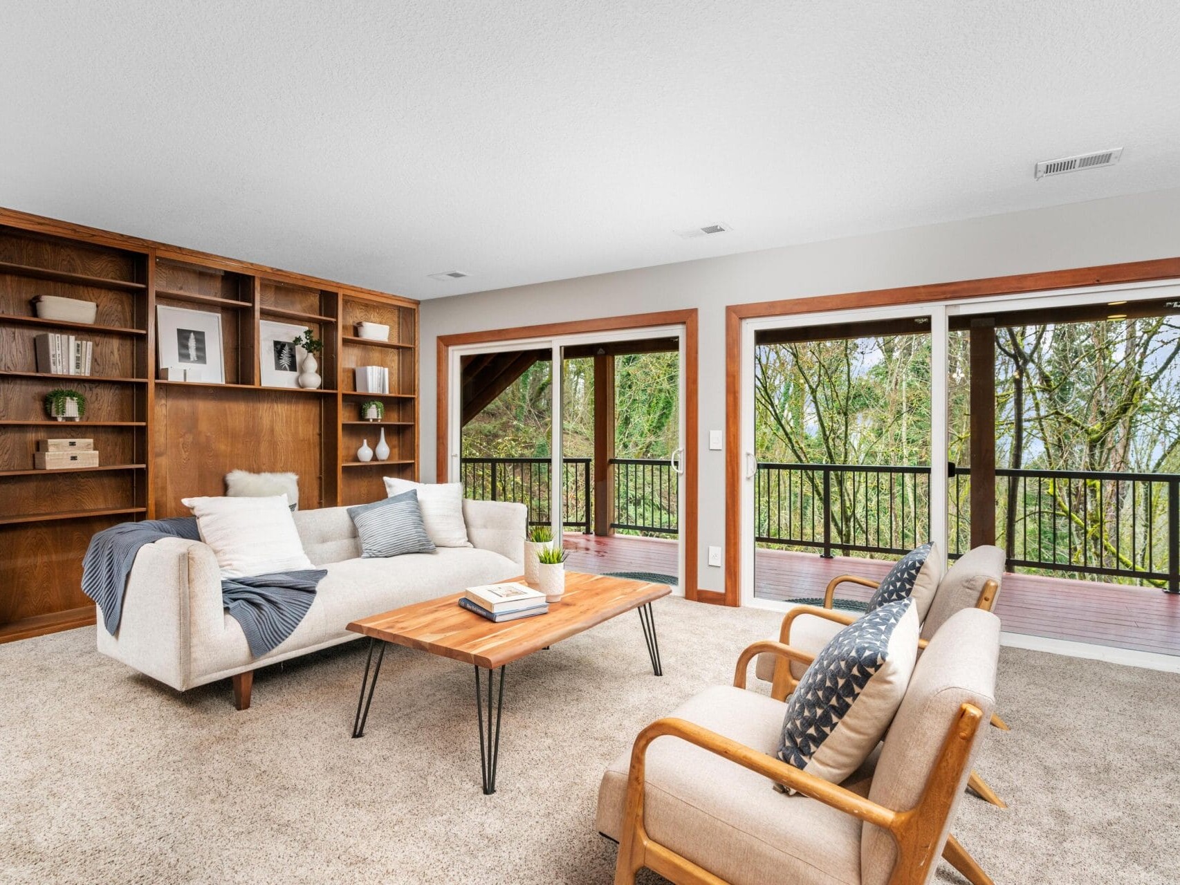 A cozy living room with carpeted floors features a wooden bookshelf, a white sofa with cushions, two cream armchairs, and a wooden coffee table. Large sliding glass doors open to a balcony with views of lush green trees.