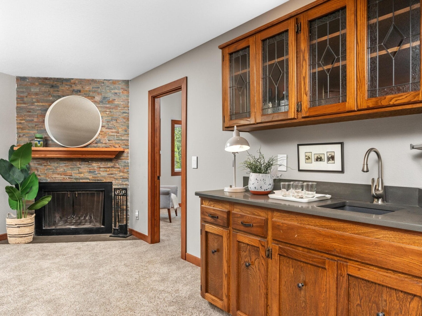 A cozy living room features a stone fireplace with a round mirror above it, a potted plant beside the hearth, and a wet bar with wooden cabinets and a countertop with a plant and glasses. An adjoining room is visible through a doorway.