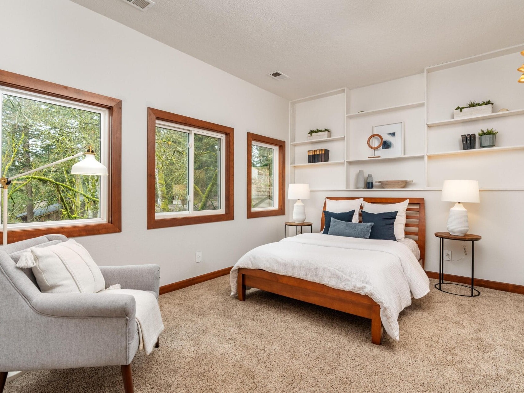 A cozy bedroom with a large bed, white bedding, and accent pillows. A gray armchair with a pillow is nearby. The room features two windows with wooden frames, a bookshelf with decor, a floor lamp, carpet, and two nightstands with lamps.