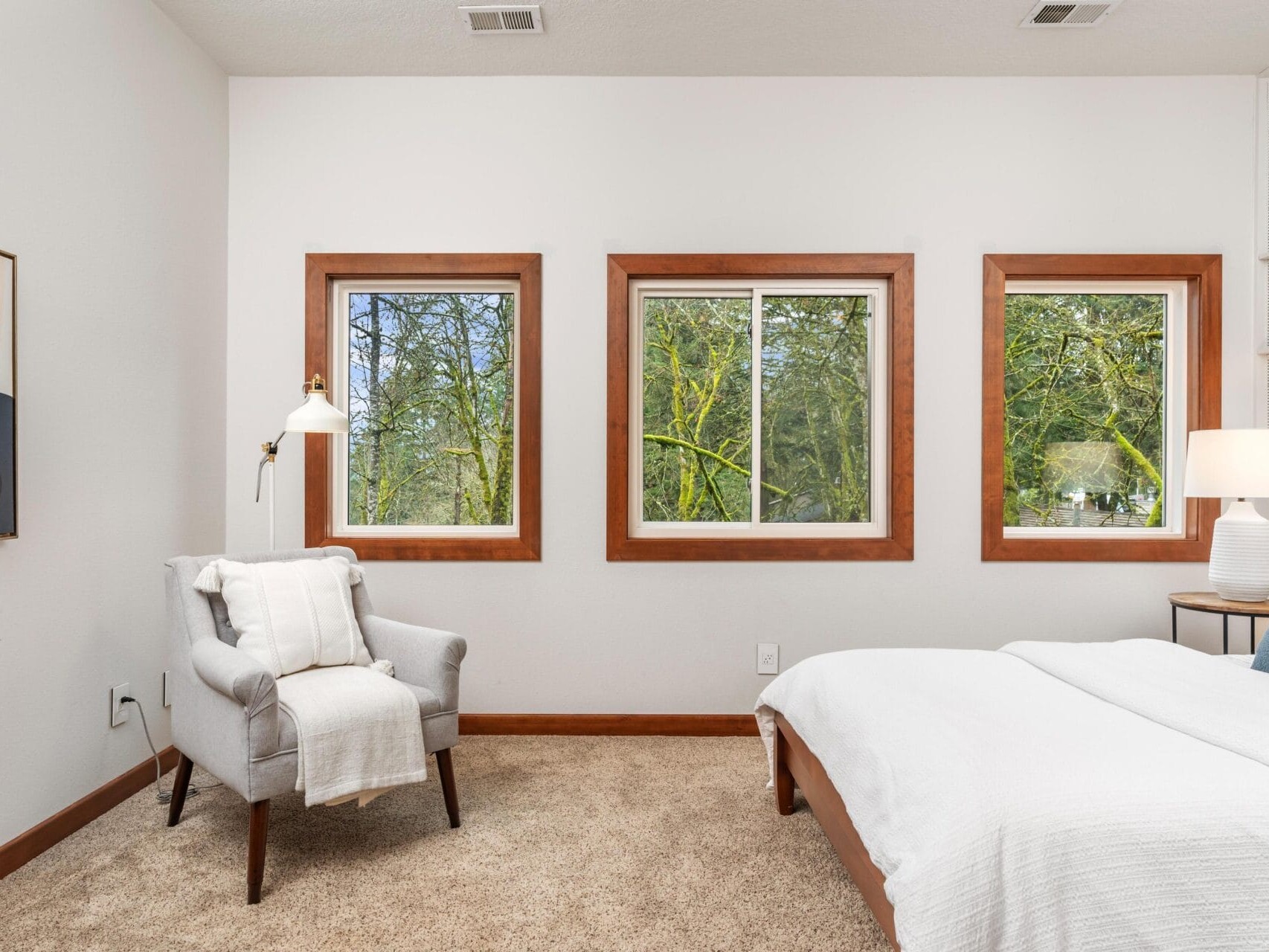 A cozy bedroom features a bed with white linens and a soft blue pillow, a gray armchair with a white blanket, and three large windows showcasing a view of trees. A modern floor lamp and a side table with a lamp complete the decor.
