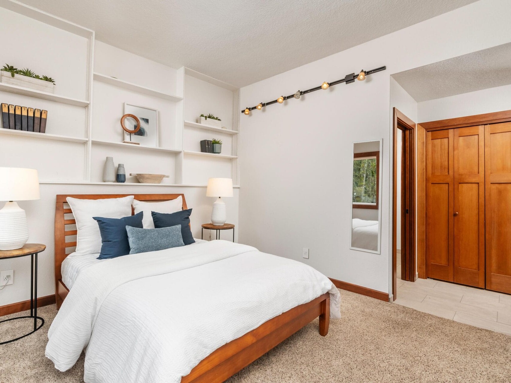 Bedroom with a wooden bed, white bedding, and blue pillows. Shelves with decor above the bed and two lamps on side tables. A string of lights is hung on the wall. Wooden closet doors are on the right, and theres a small mirror beside them.