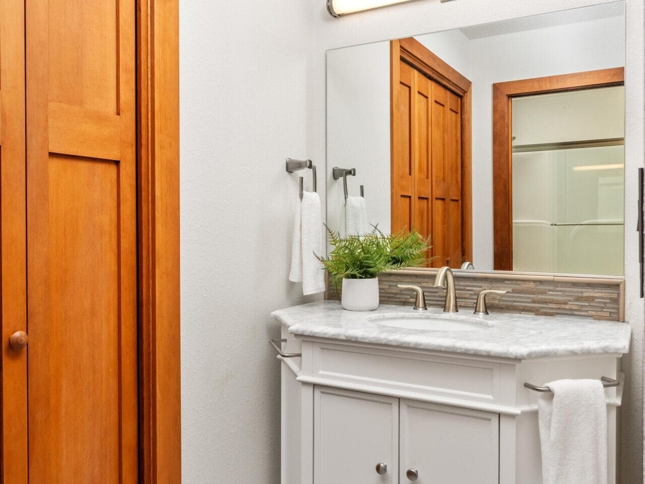 A bathroom featuring a wooden sliding door, a white vanity with a marble countertop, dual faucets, and a large rectangular mirror. Theres a bright overhead light and a small plant on the counter. Towels hang on the rack and door.