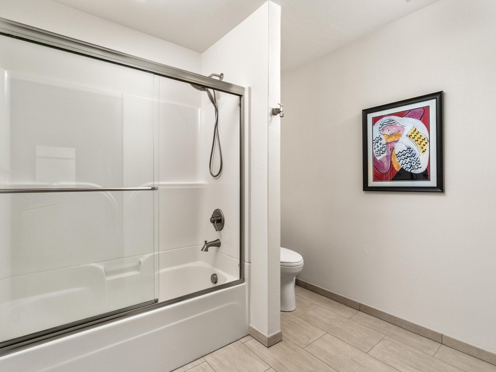 Modern bathroom with a glass-enclosed bathtub and sliding doors on the left. A toilet is partially visible behind the wall. A framed abstract painting with vibrant colors hangs on the right wall. The floor is tiled in a neutral tone.