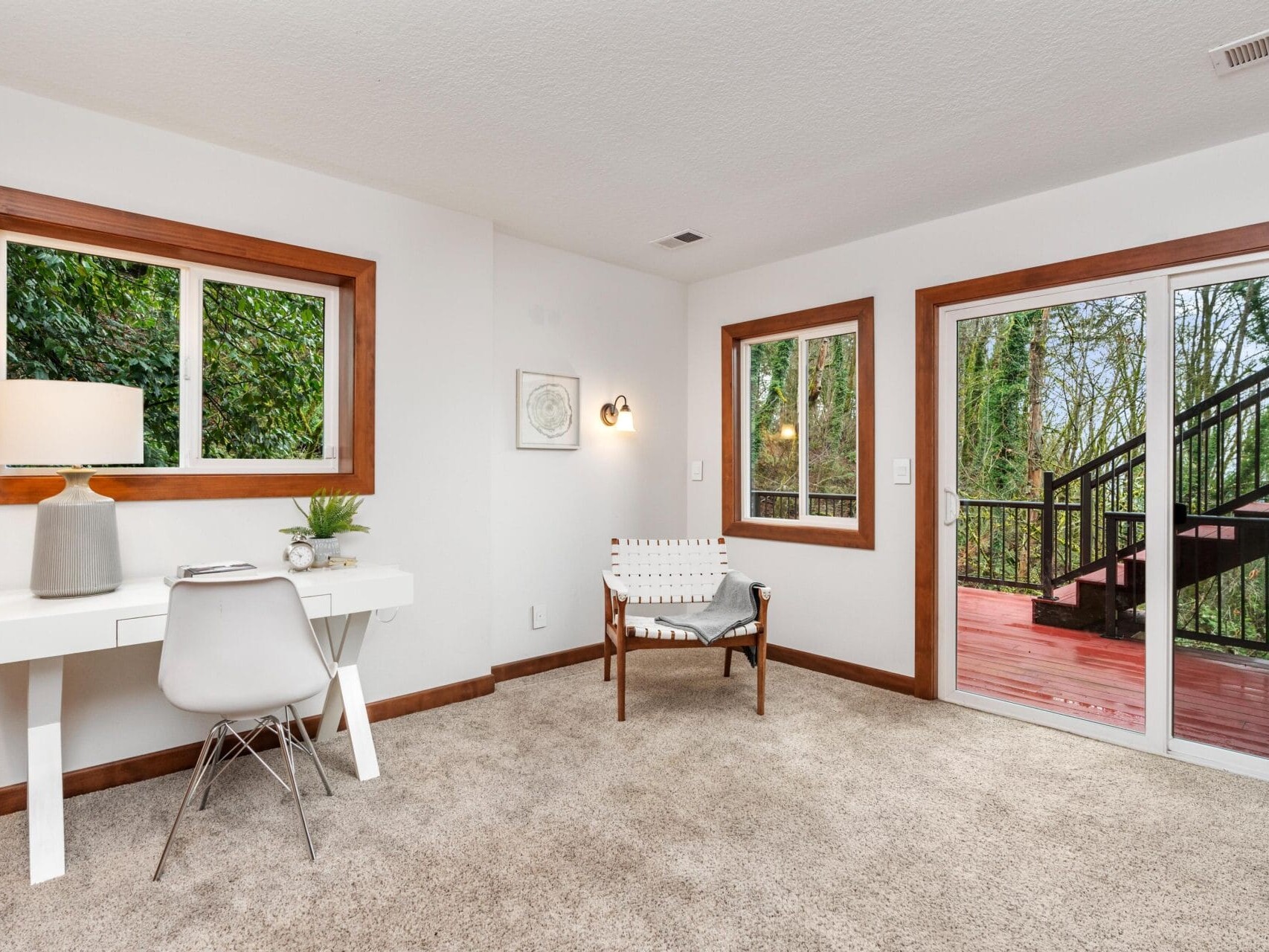 A bright room with beige carpet, featuring a white desk with a lamp and chair, a wooden chair, and large windowed doors leading to a red wooden deck. The windows and doors offer a view of greenery outside.