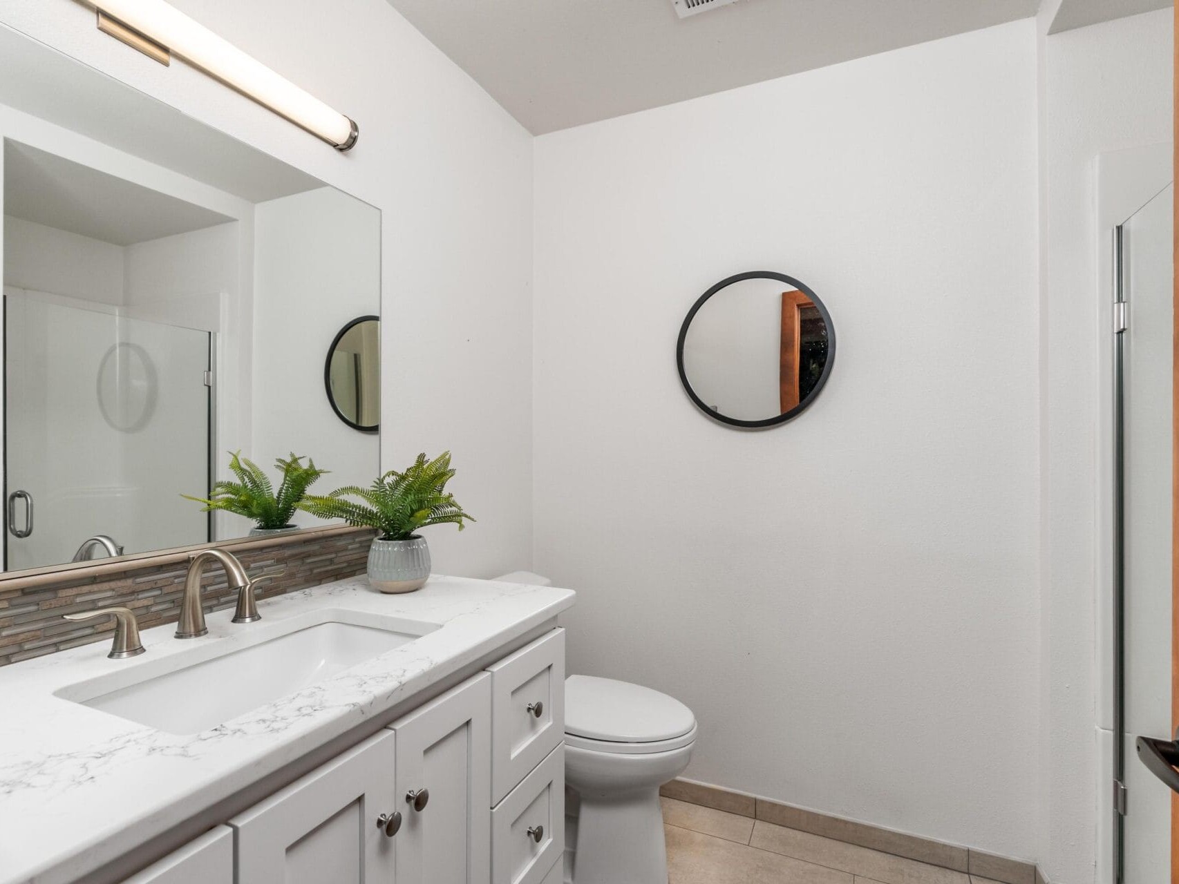 A modern bathroom with a white countertop and sink, silver faucet, and potted fern. Theres a large rectangular mirror above, a round mirror on the opposite wall, and a glass shower partially visible. The room is well-lit and minimalistic.