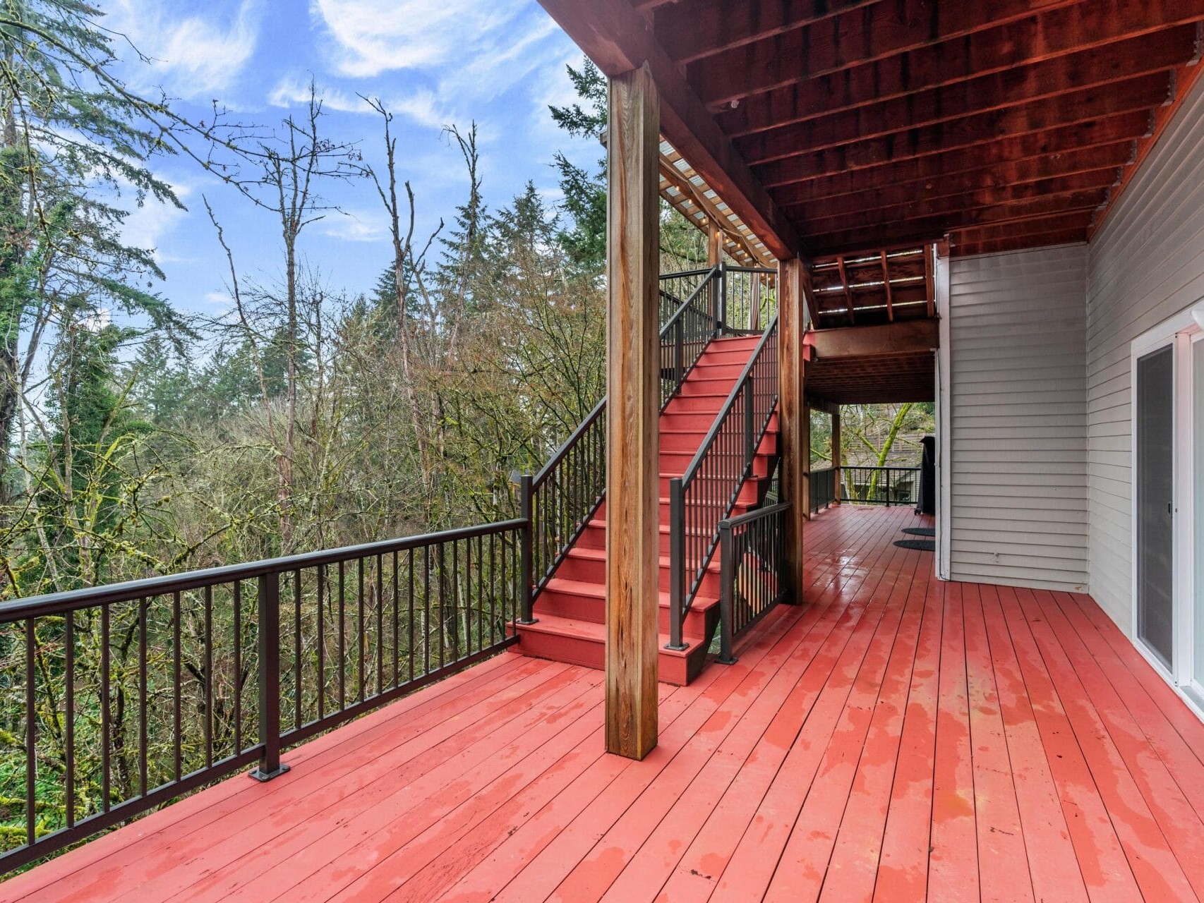 A wooden deck with red flooring and black railings, featuring a staircase leading up. Surrounded by tall trees under a blue sky with wispy clouds, the area is next to a light gray building with large sliding glass doors.