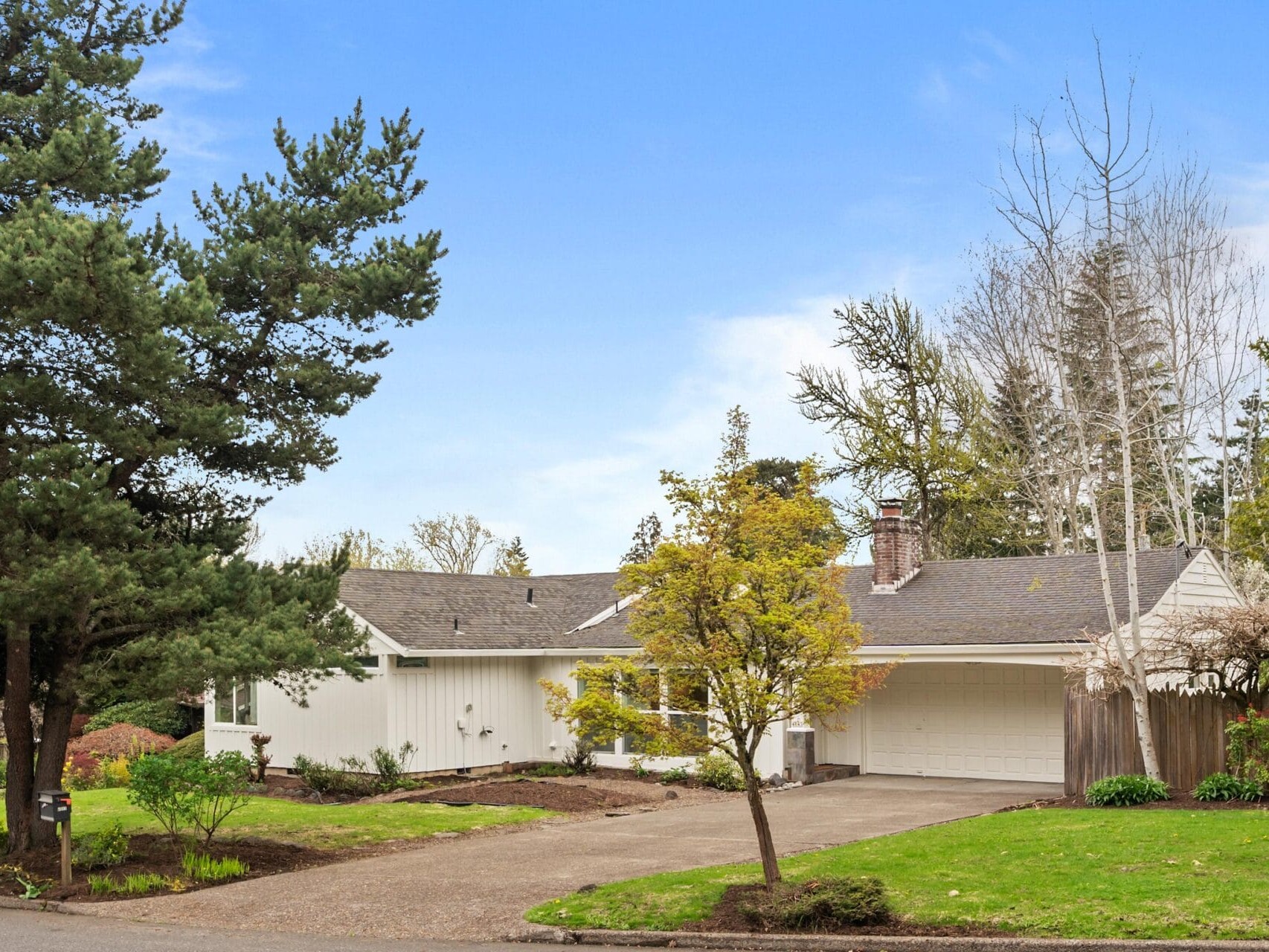 A single-story white house with a gray roof and attached two-car garage sits on a quiet suburban street, surrounded by trees, shrubs, and a well-maintained lawn under a blue sky.