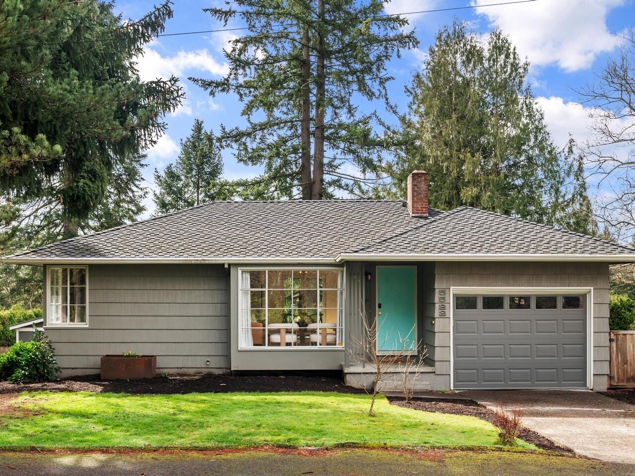 Single-story gray house with a gable roof, large window, and green front door. Attached one-car garage to the right. Surrounded by lush trees and a green lawn under a partly cloudy sky.