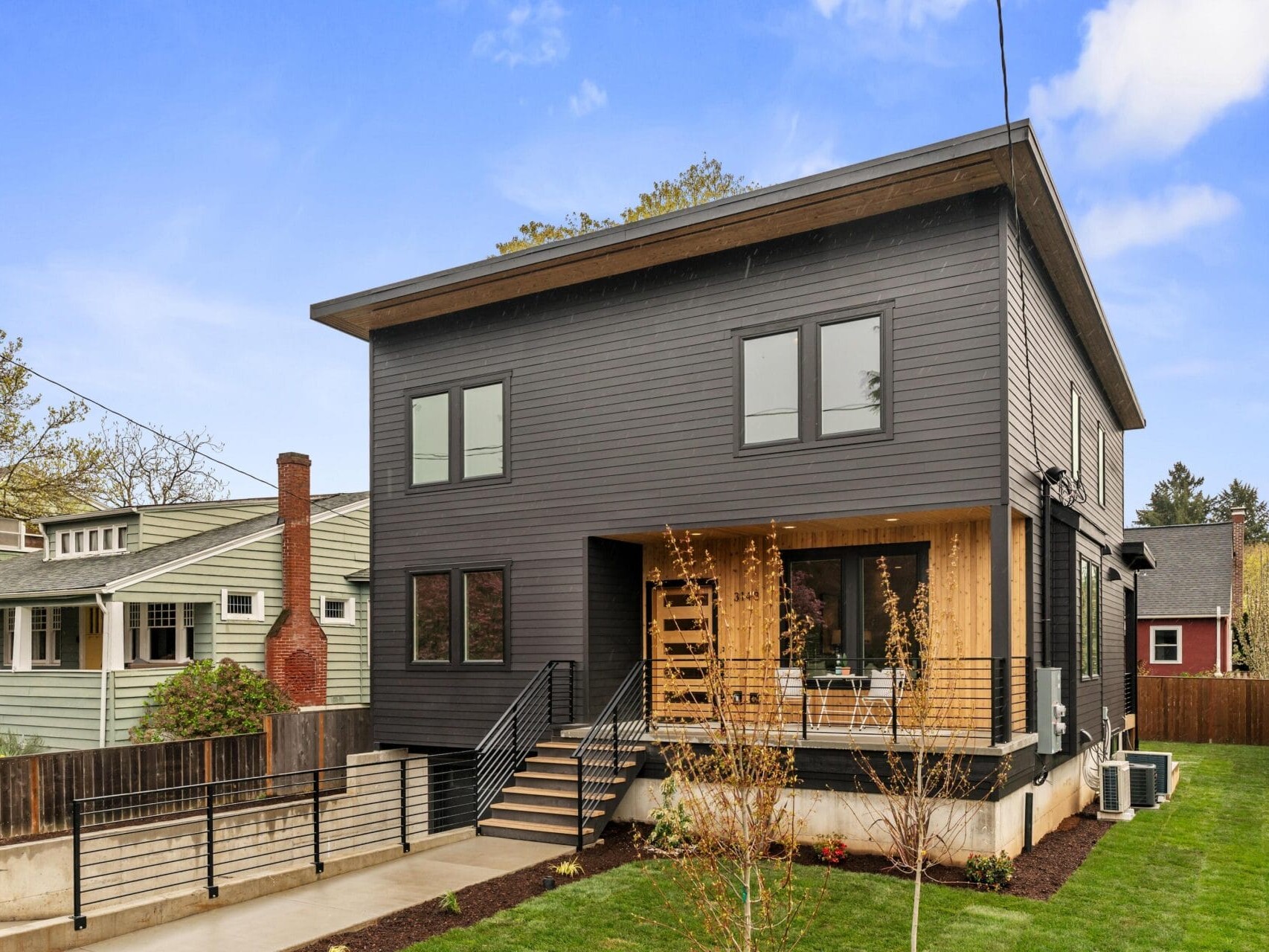 Modern two-story house with dark gray siding and large windows. A small front porch has a wooden facade and metal railings. A manicured lawn and driveway lead to the entrance. Neighboring homes and trees visible under a blue sky.